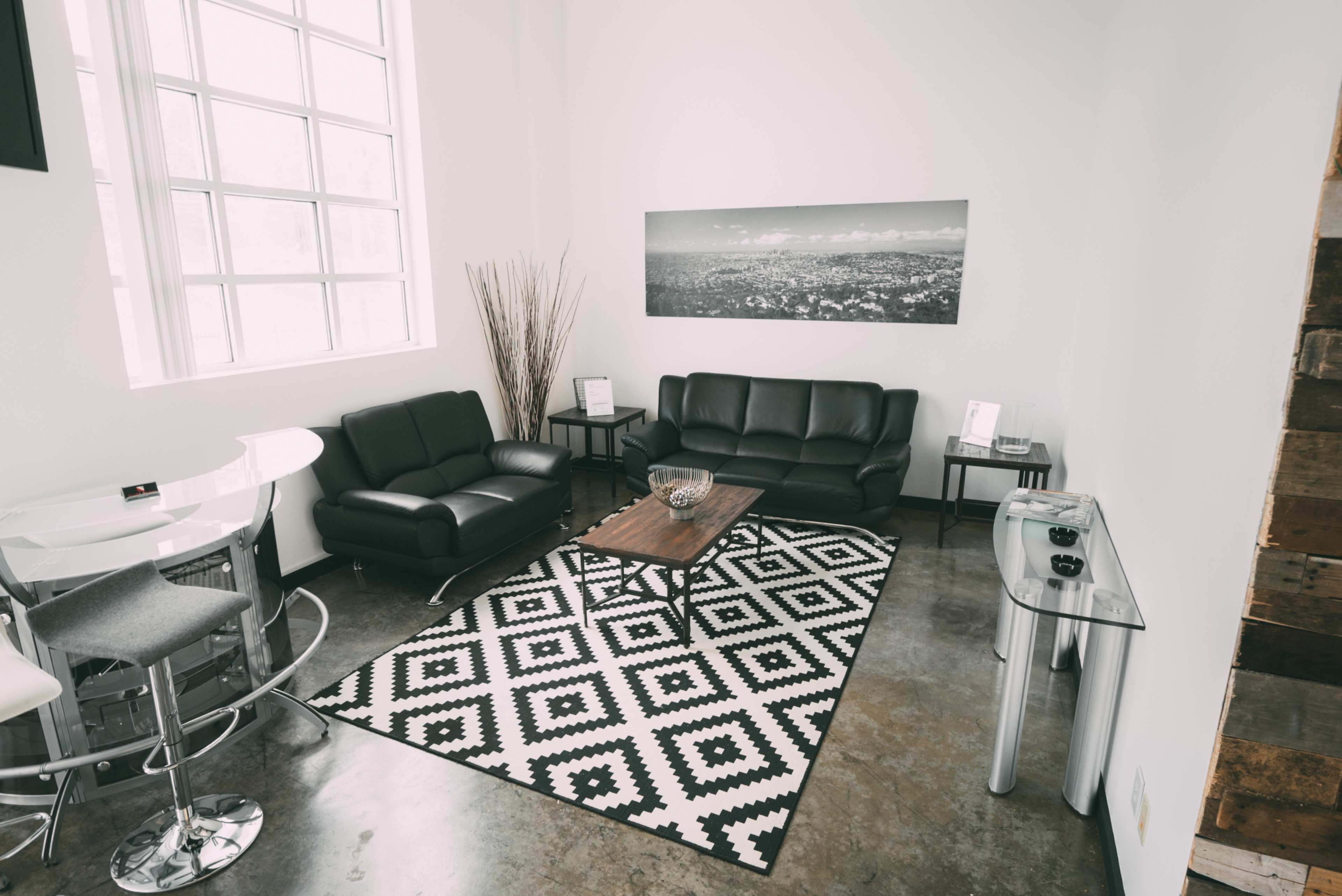 A modern living area with black leather sofas, a wooden coffee table on a patterned rug, and large windows allowing natural light.