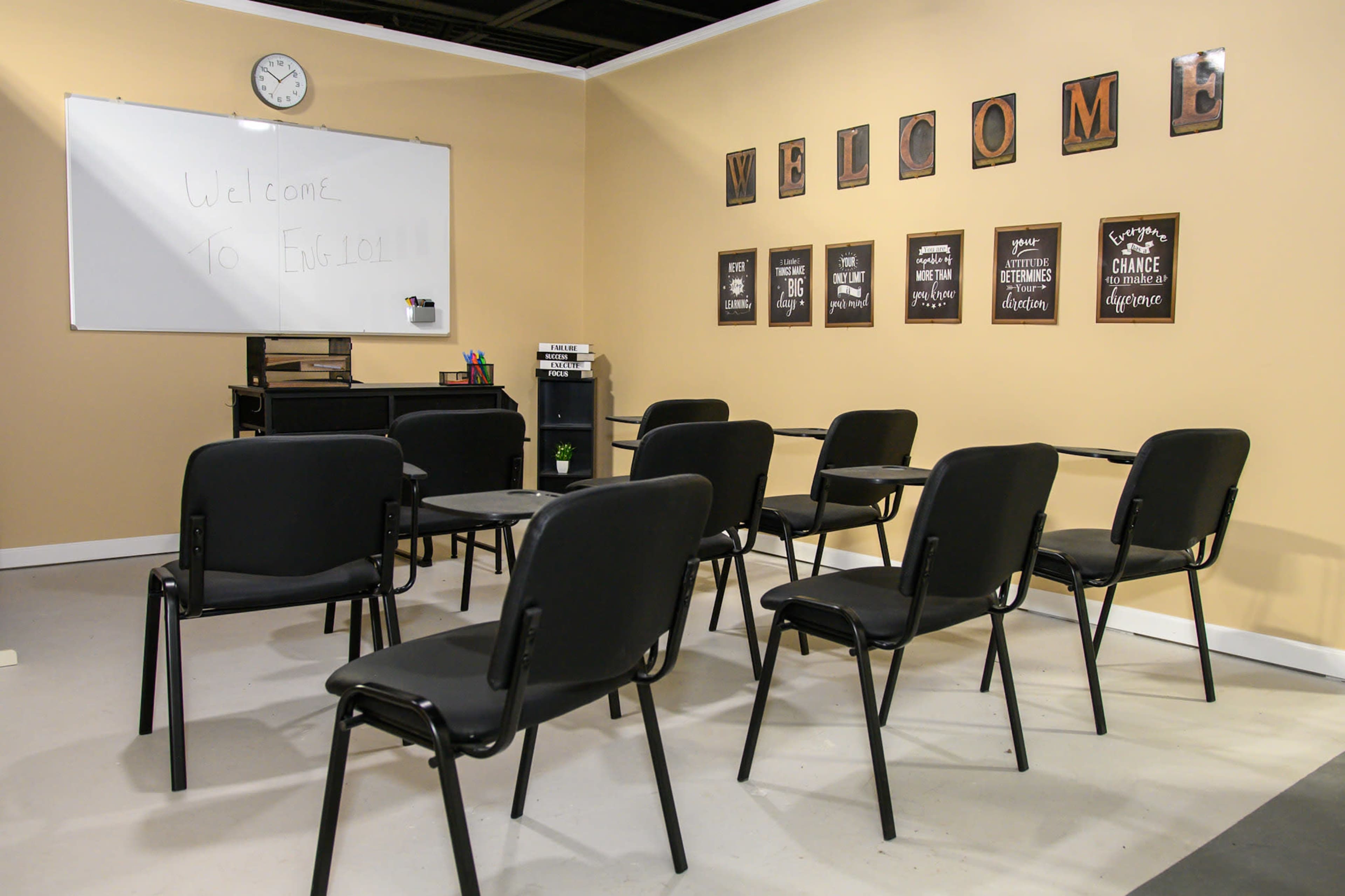 A classroom setting is depicted, featuring rows of black chairs facing a whiteboard with a clock on the wall and motivational posters arranged above.