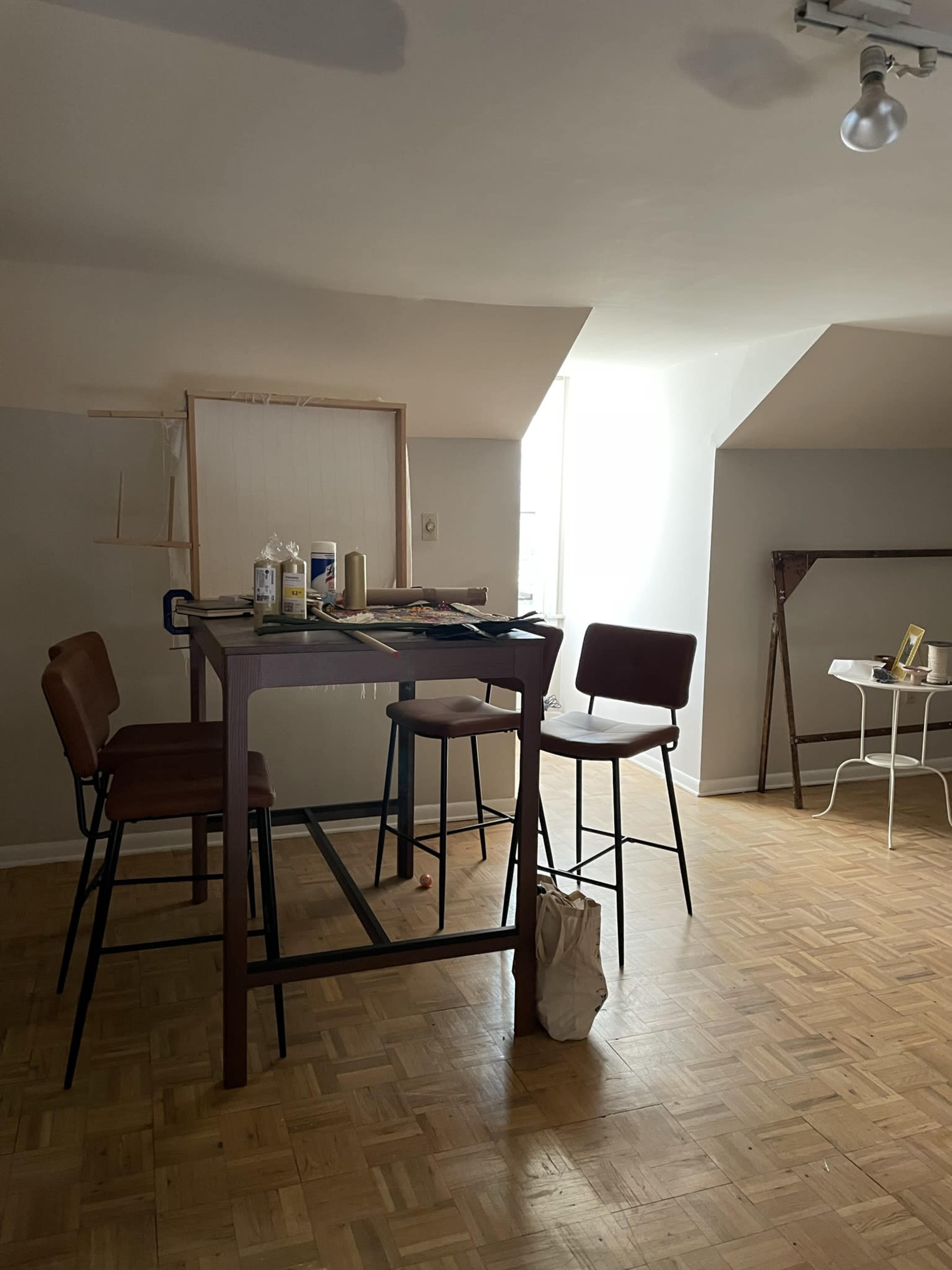 A spacious room with a high table and three chairs, illuminated by natural light from a window, along with an easel and a small table in the background.
