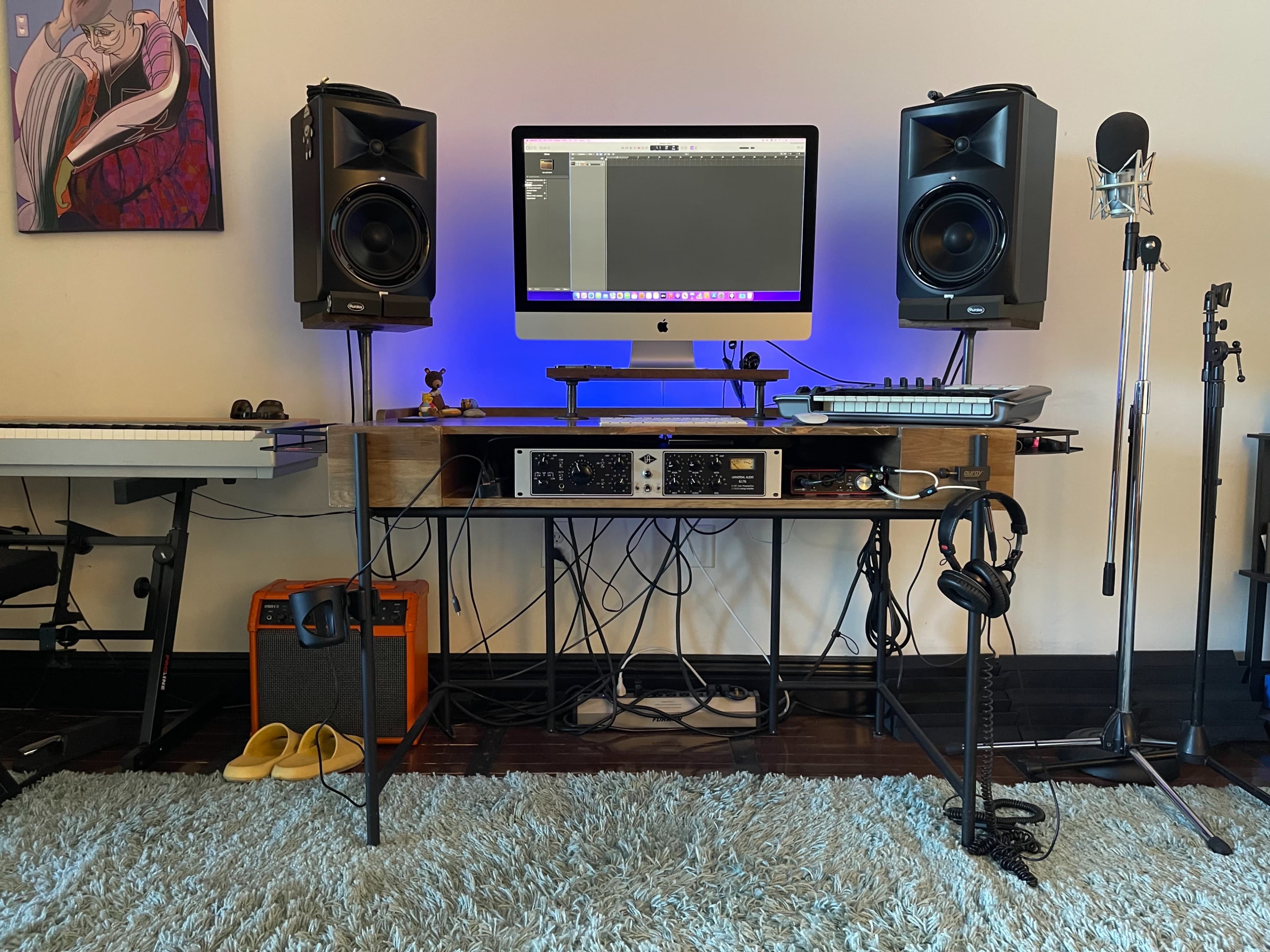 A music production setup features a computer monitor, studio monitors, a microphone, and various musical equipment on a wooden desk with headphones and an amplifier on a light-colored rug.