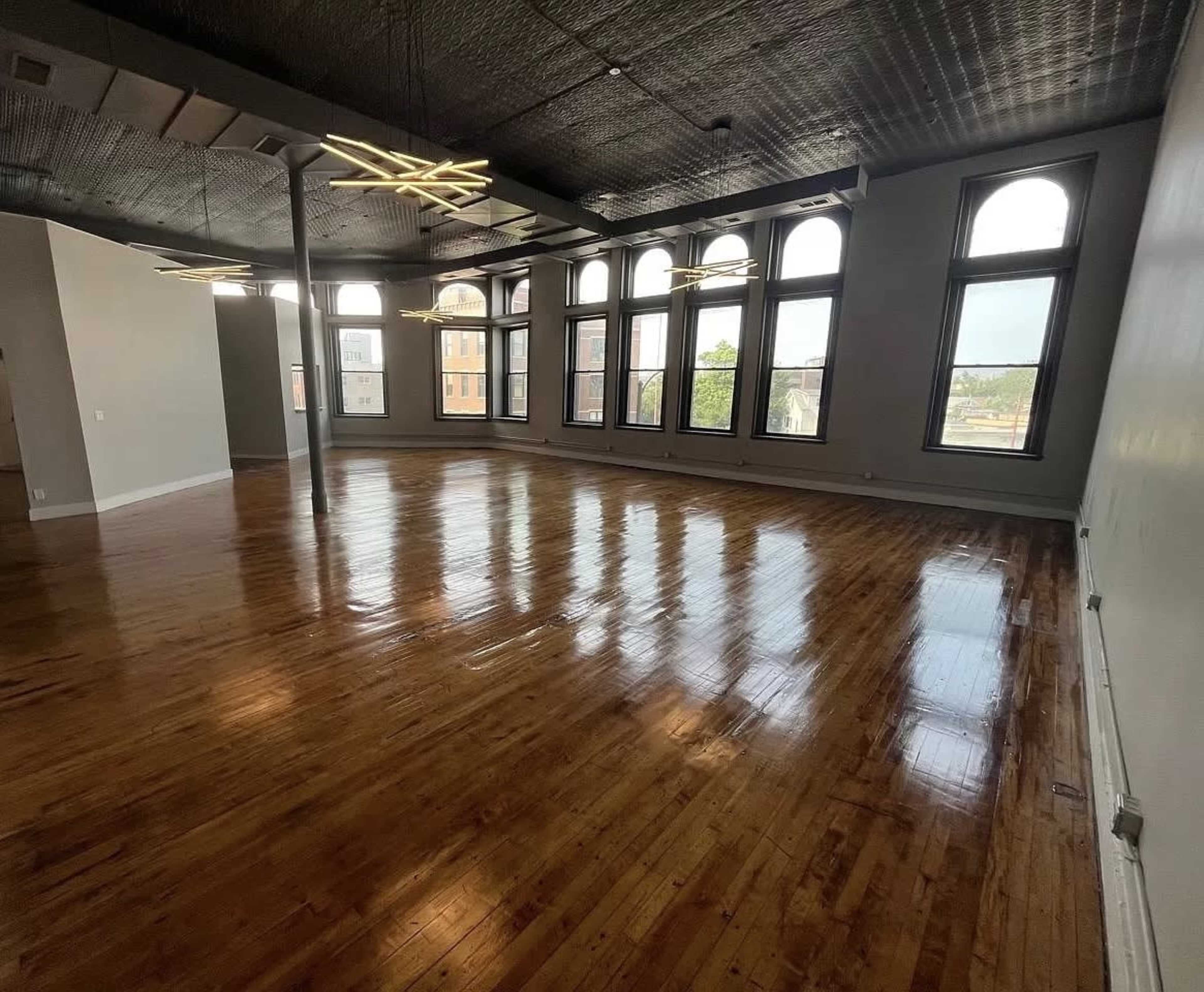 The image shows a spacious, empty room with polished wooden floors and large arched windows that let in natural light.