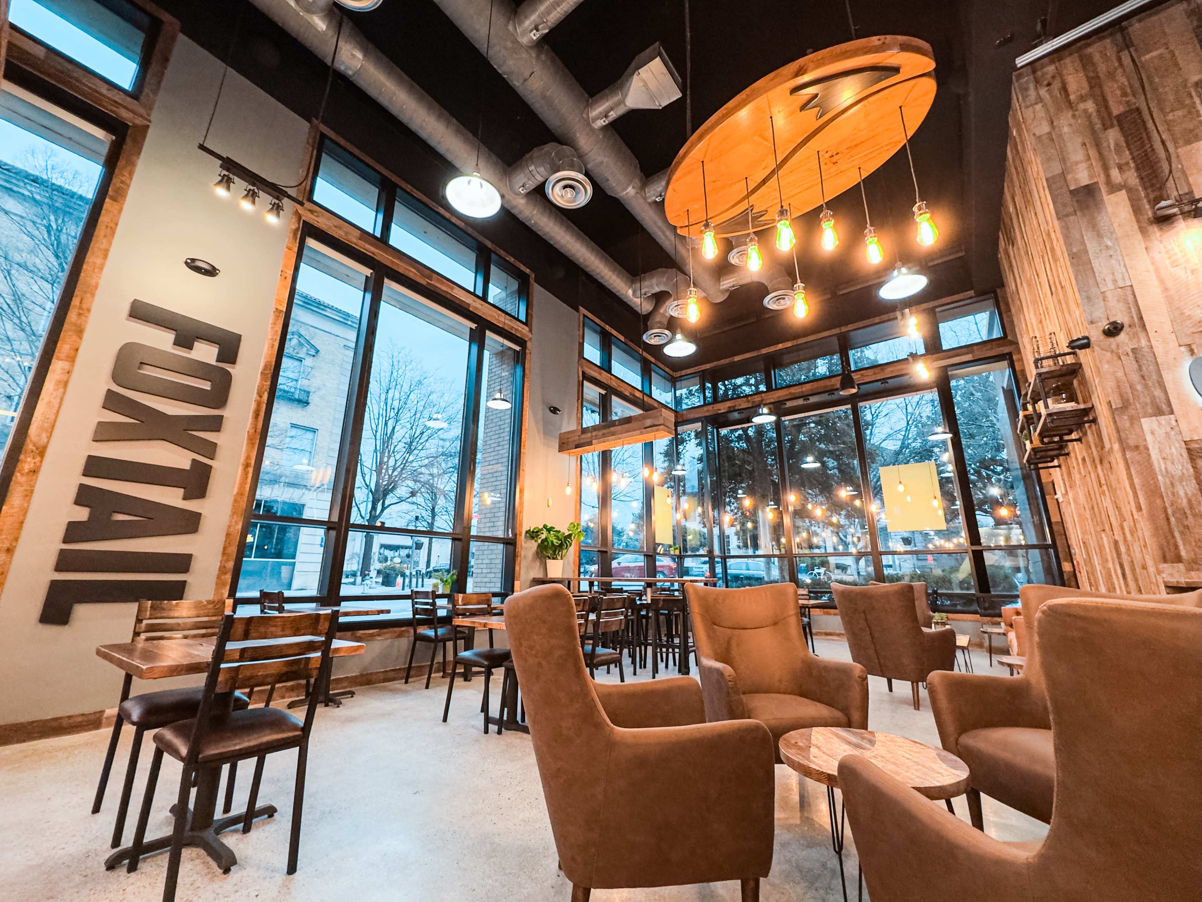 The image shows a modern café interior with wooden accents, large windows, and a variety of seating arrangements, including plush armchairs and tables.
