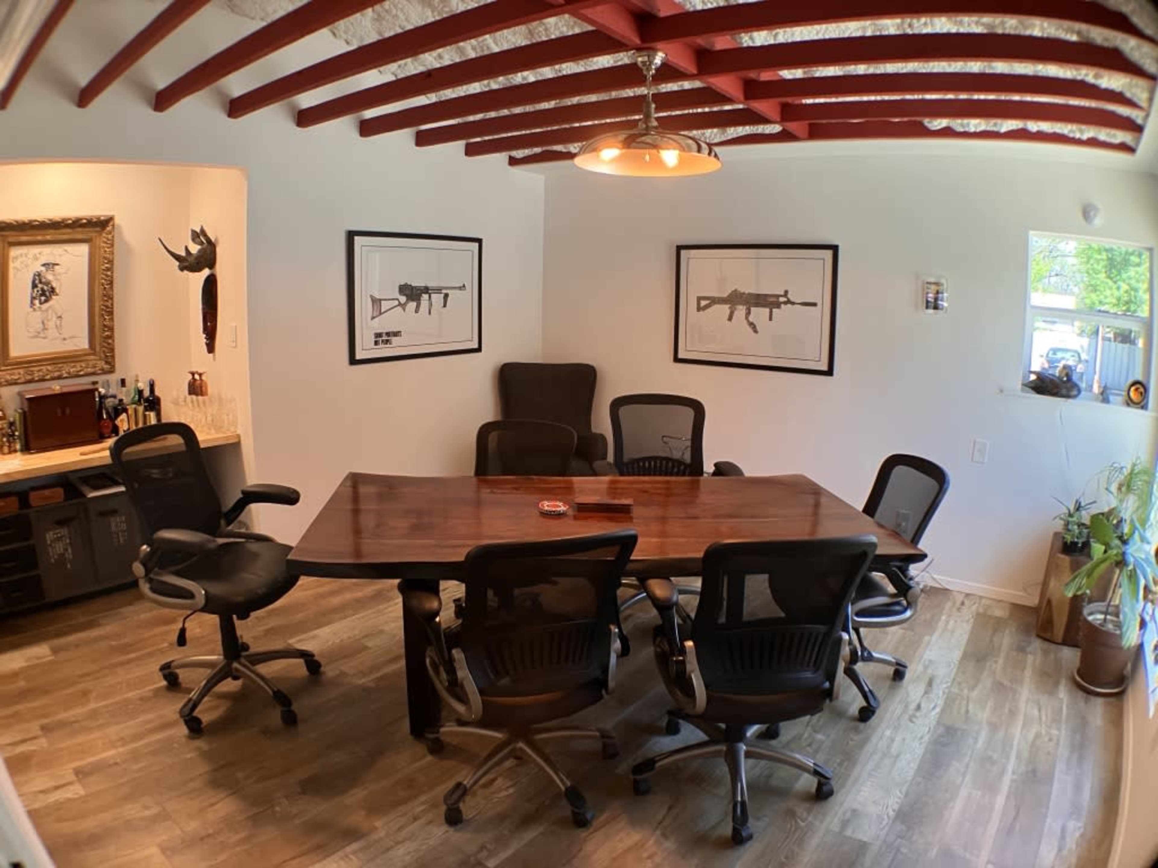 A conference room features a long wooden table surrounded by several black office chairs, with framed artwork on the walls and a window with natural light.