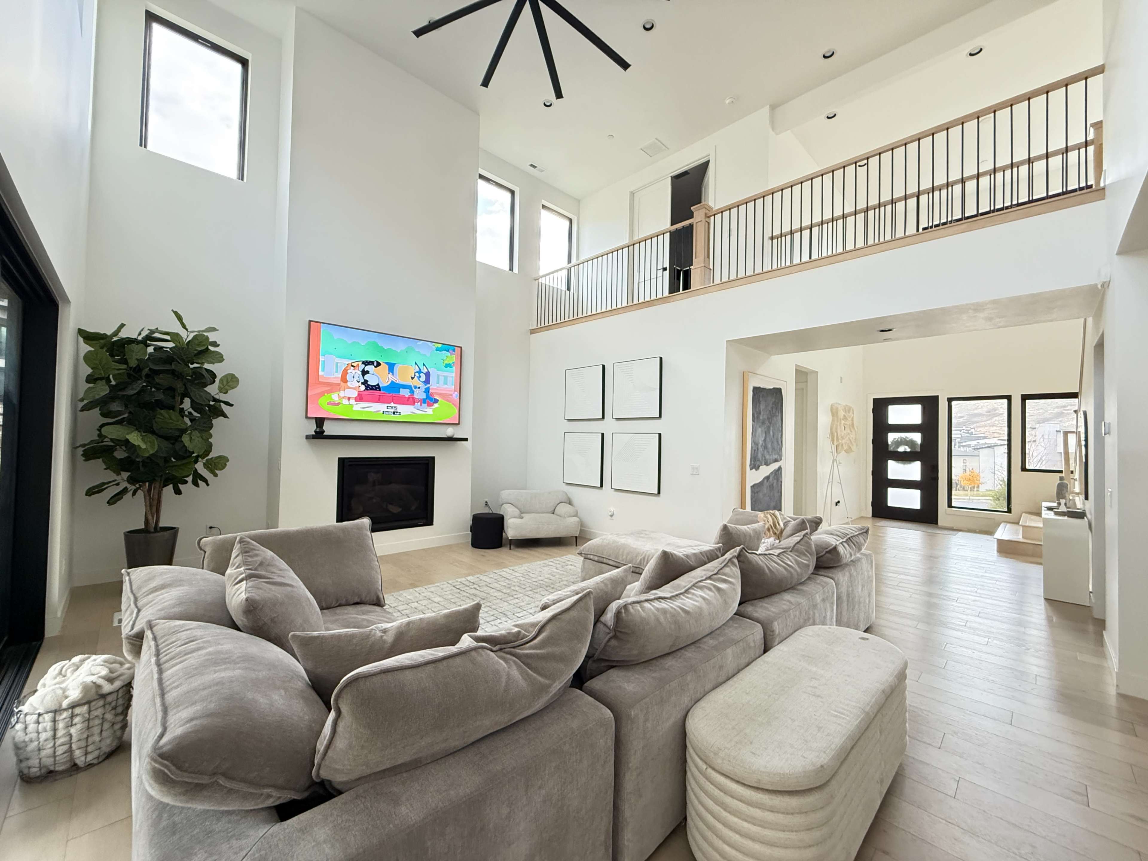 The image shows a spacious modern living room with high ceilings, a large sectional sofa, a wall-mounted television displaying colorful animations, and an open view towards a front entryway.