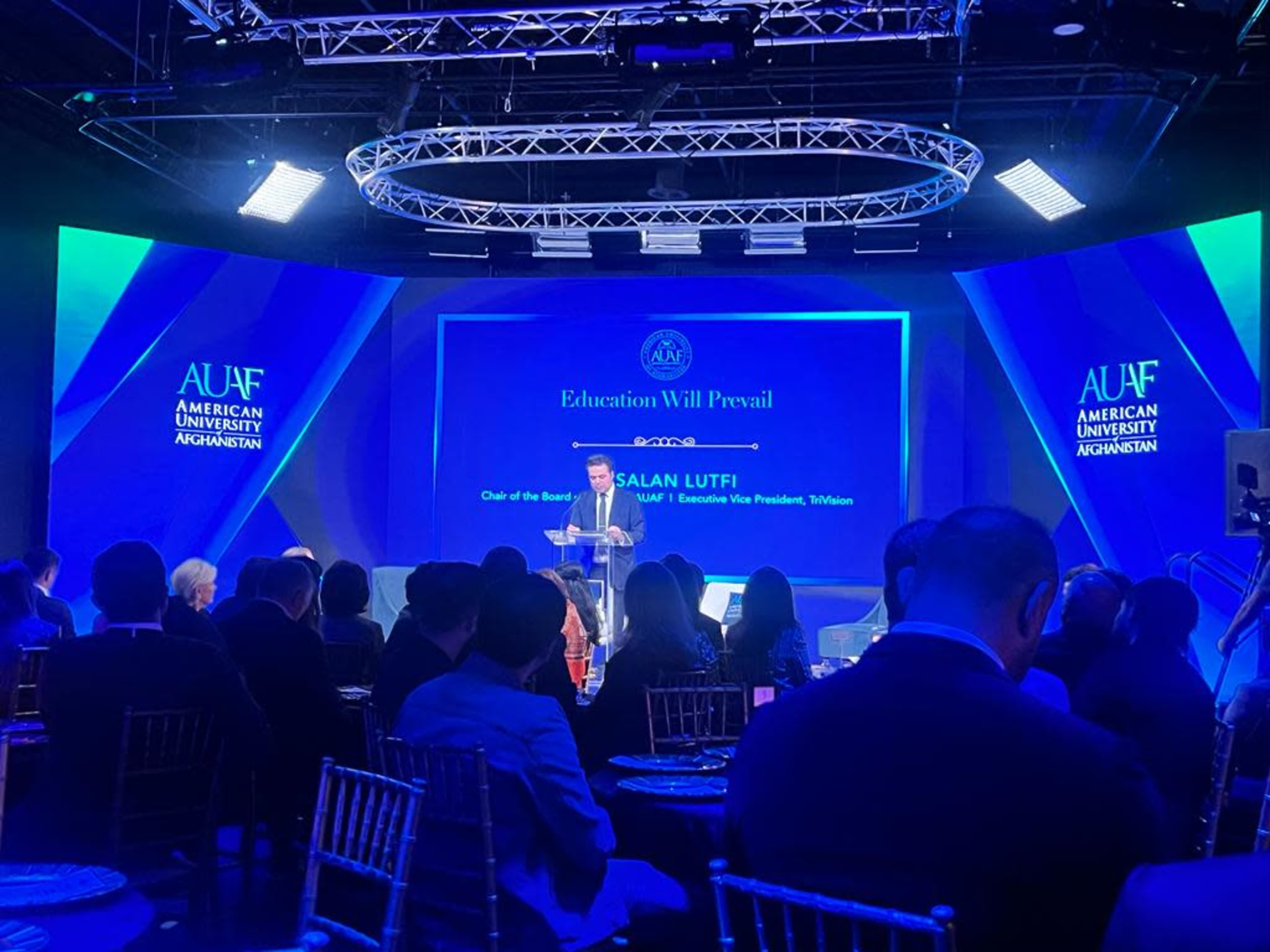 A speaker stands at a podium during an event at the American University of Afghanistan, with a blue and white backdrop featuring the university's logo and lighting.