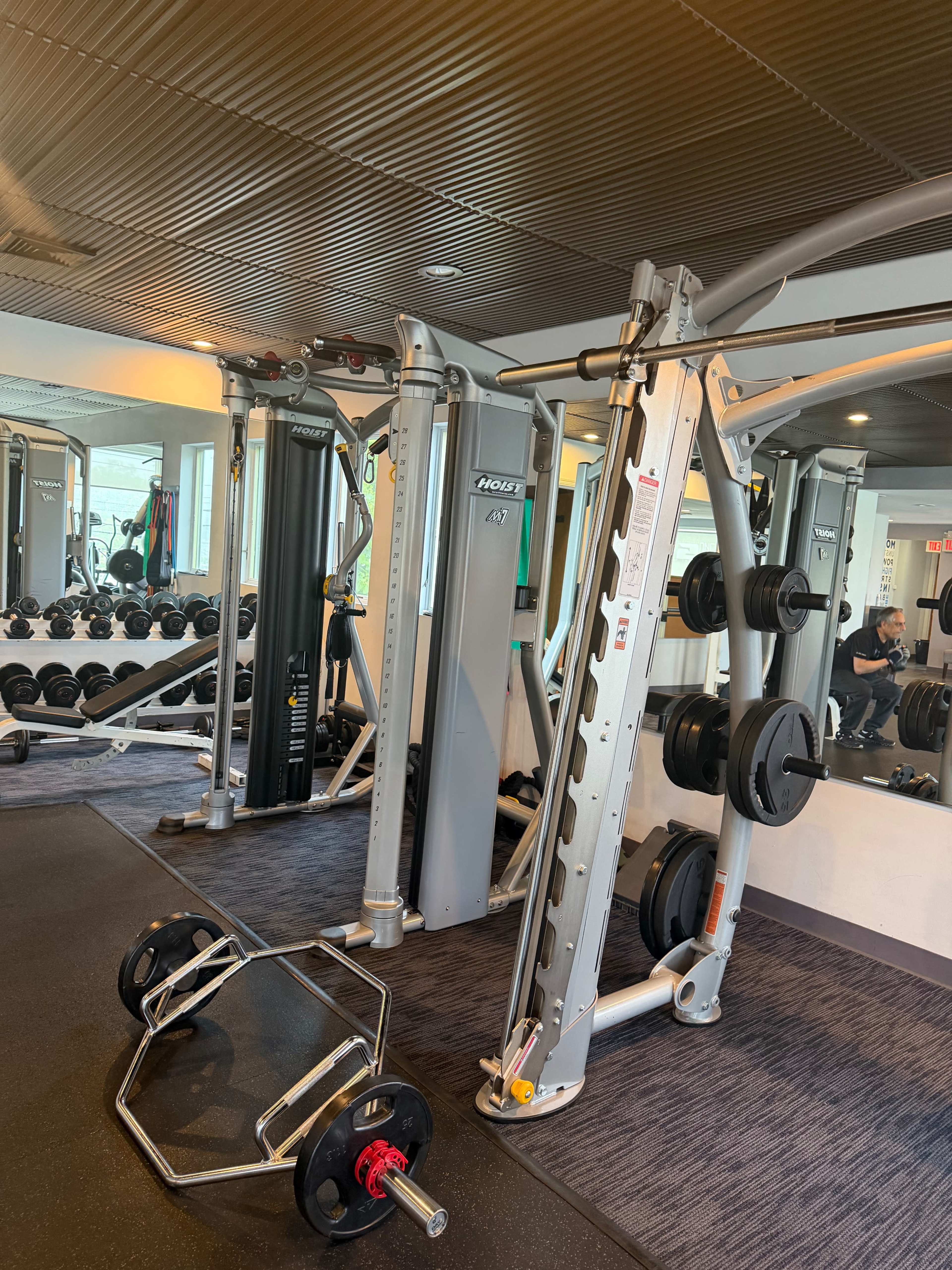 The image shows a modern gym interior with weightlifting equipment, including a cable machine and barbell, and weight plates loaded on a nearby rack.