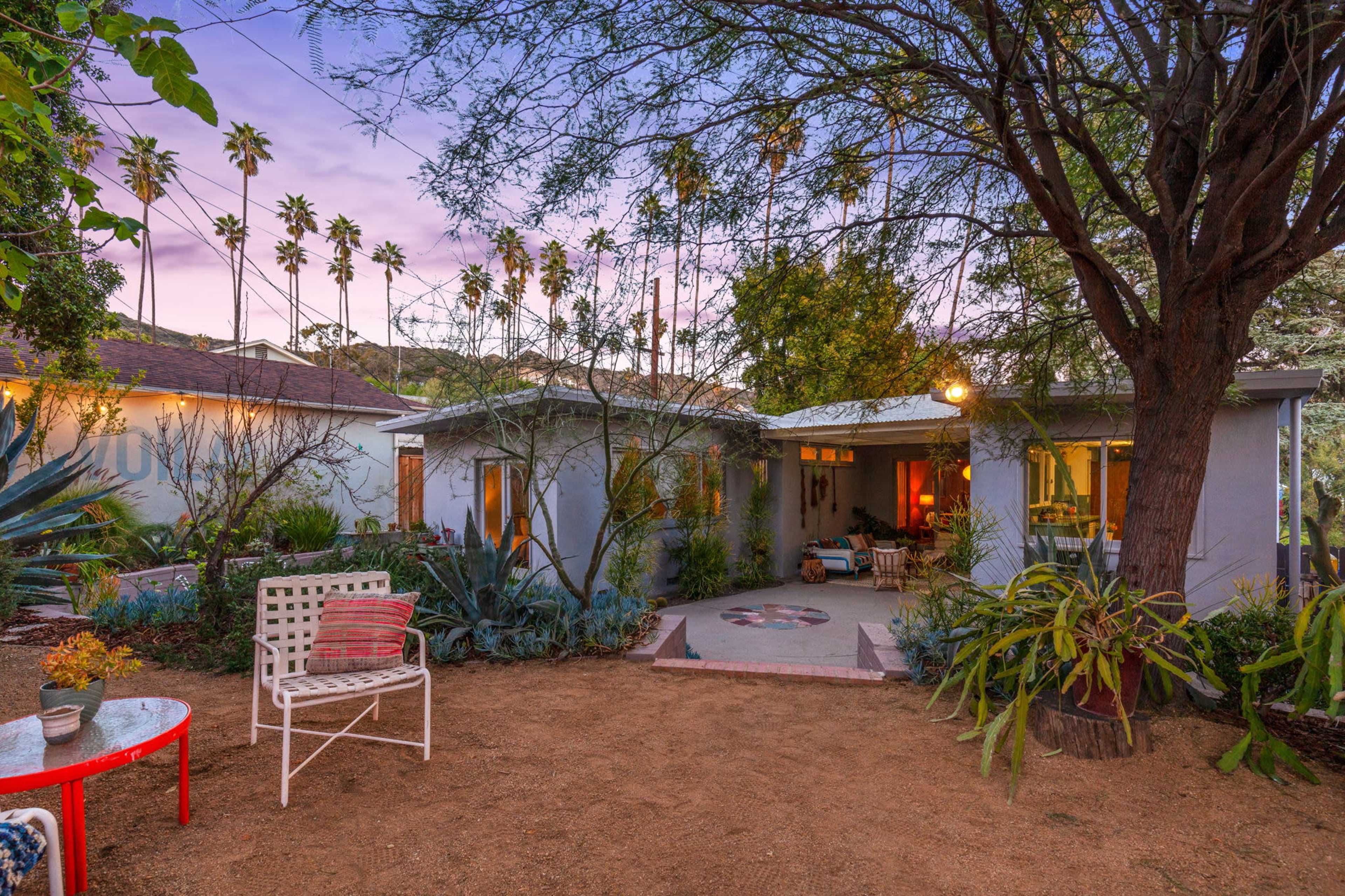 A small, modern home with large windows is surrounded by trees and plants, featuring a cozy outdoor seating area and a gravel path.