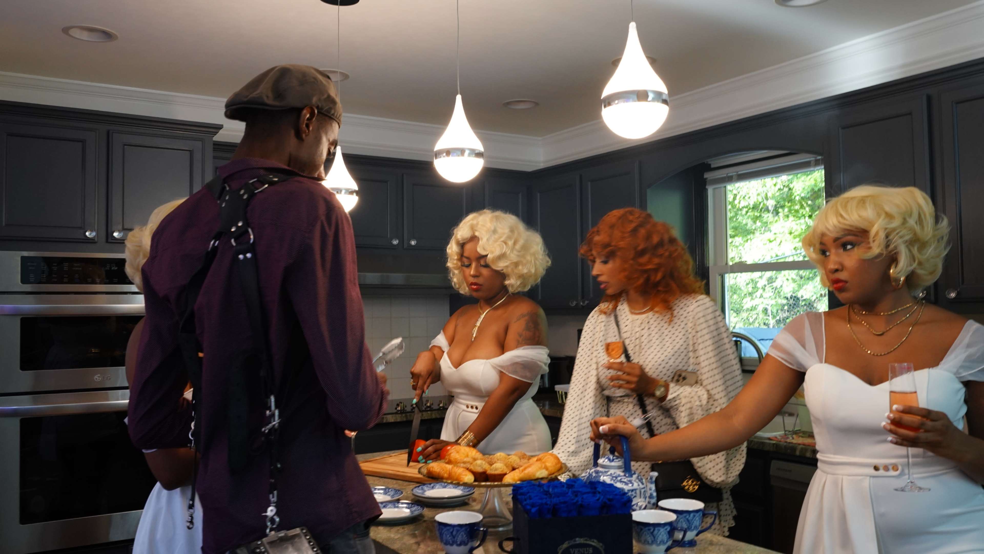 A group of four people, two with blonde curly hairstyles and two with orange hair, gather in a kitchen with gray cabinets, discussing food on a countertop.
