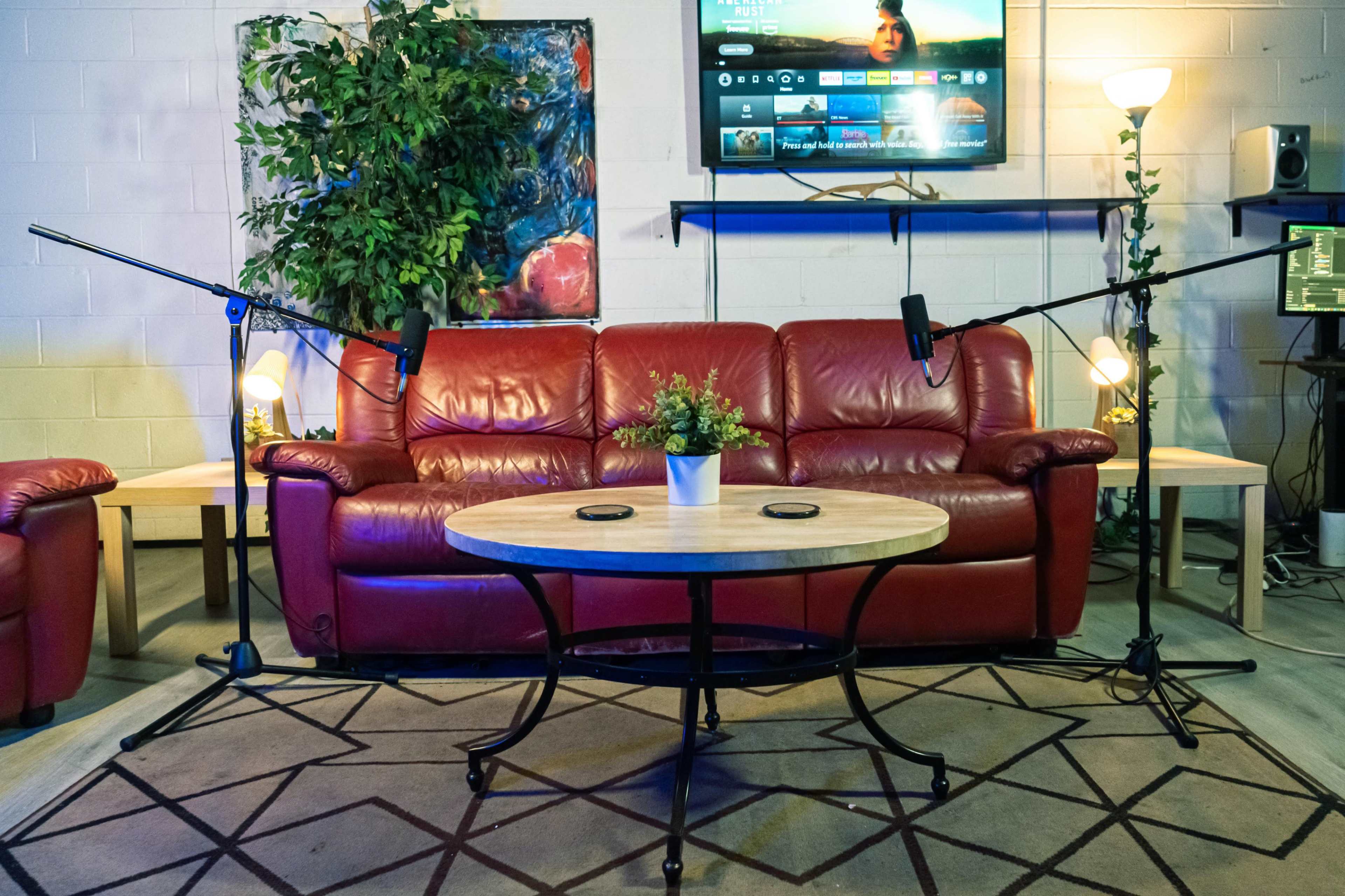 The scene features a red leather couch with two microphones on stands, a circular wooden coffee table with a small plant, and a television mounted on the wall above.