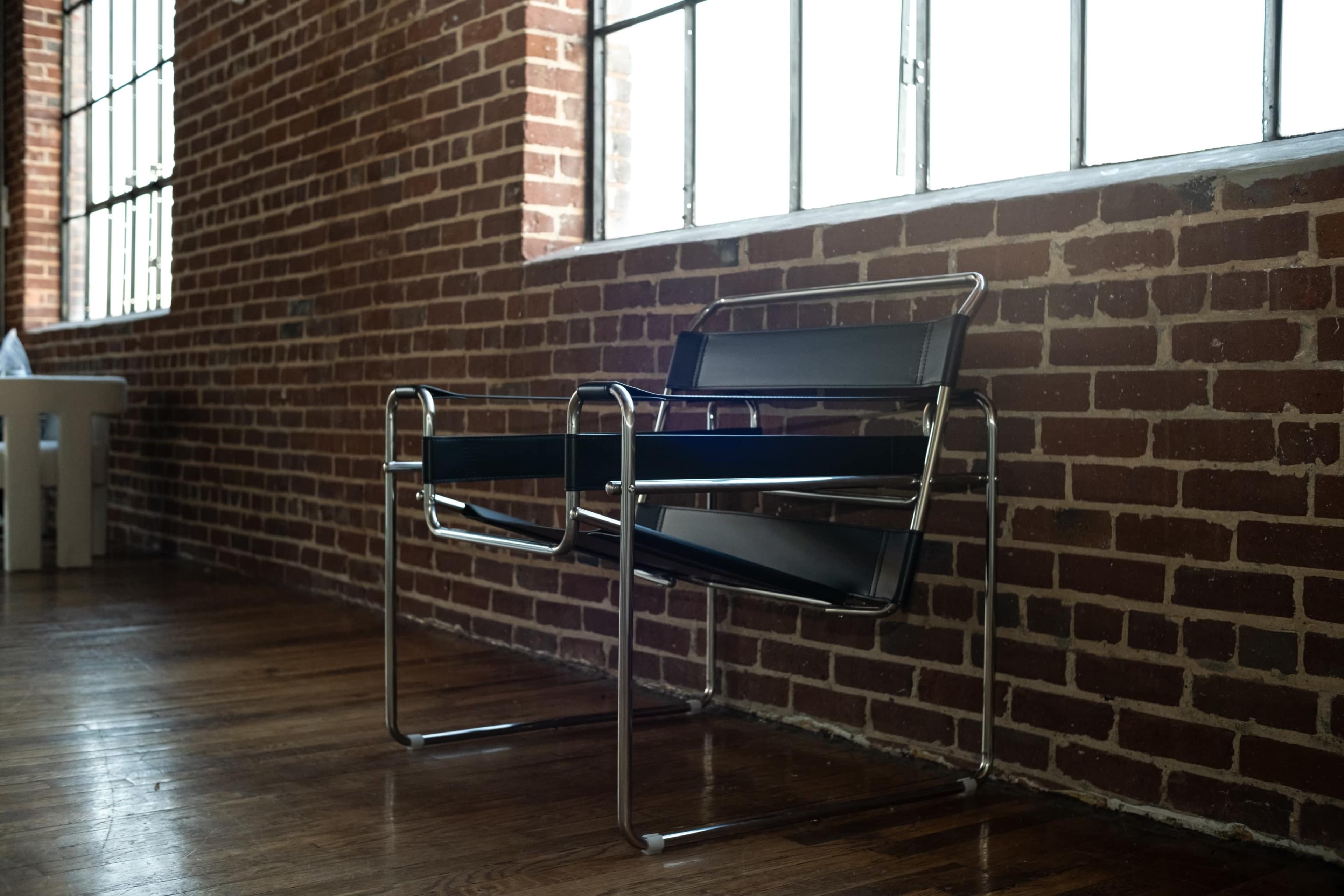 A metal-framed chair with black straps is positioned beside a large window in a brick-walled interior.