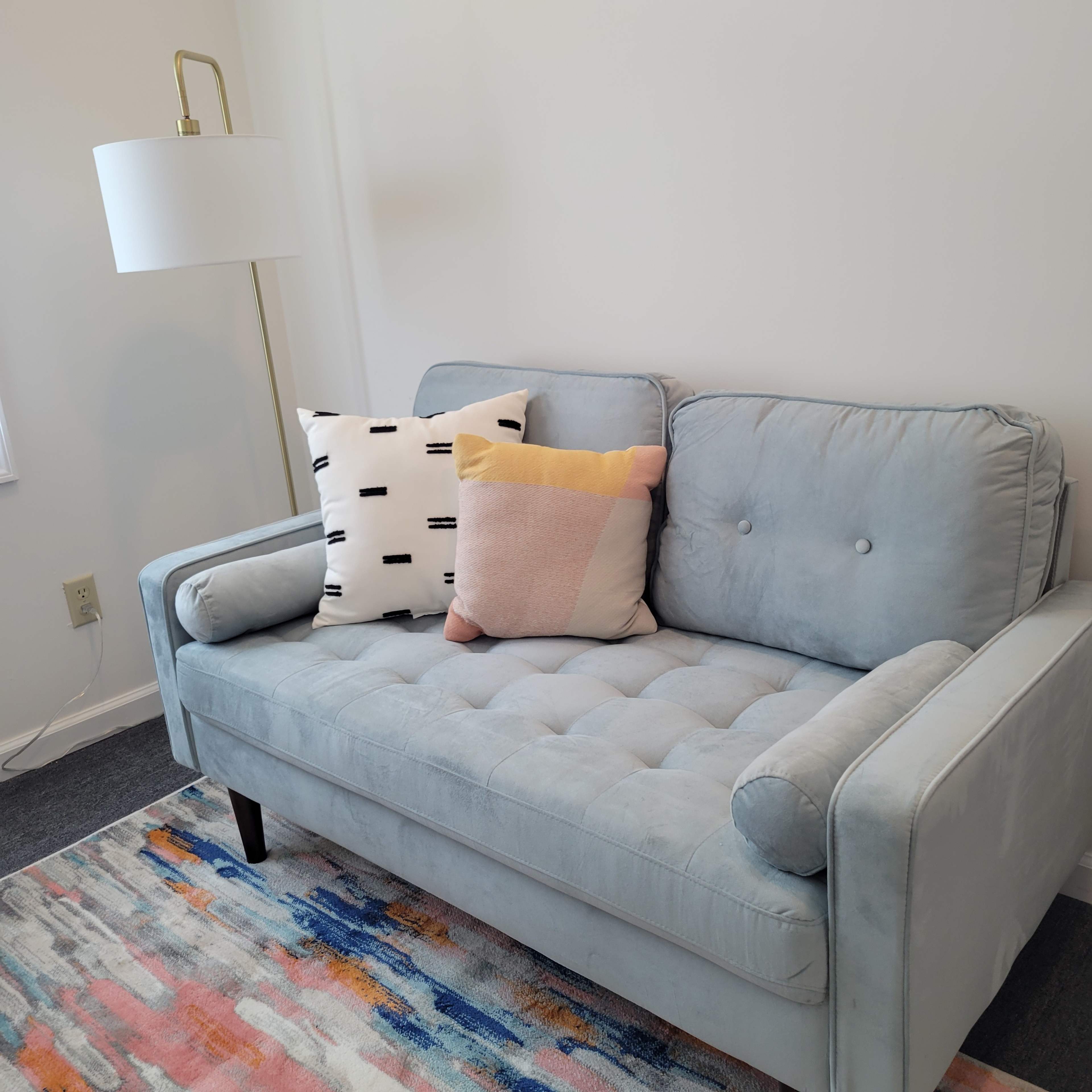 A light blue sofa with decorative pillows sits on a colorful rug next to a tall lamp in a bright room.