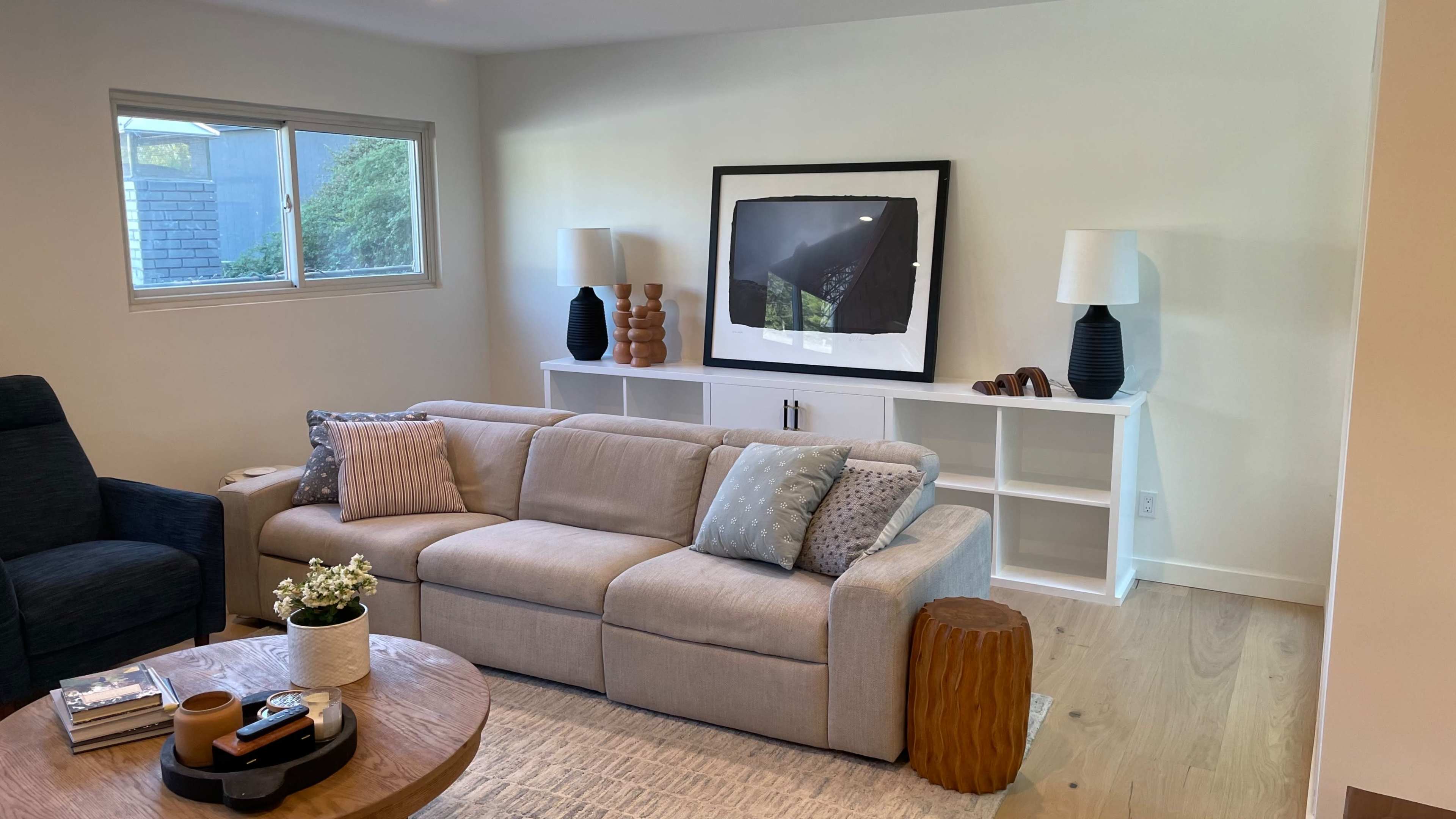 A modern living room with a beige couch, a round wooden coffee table, and a white console table adorned with decorative lamps and artwork.
