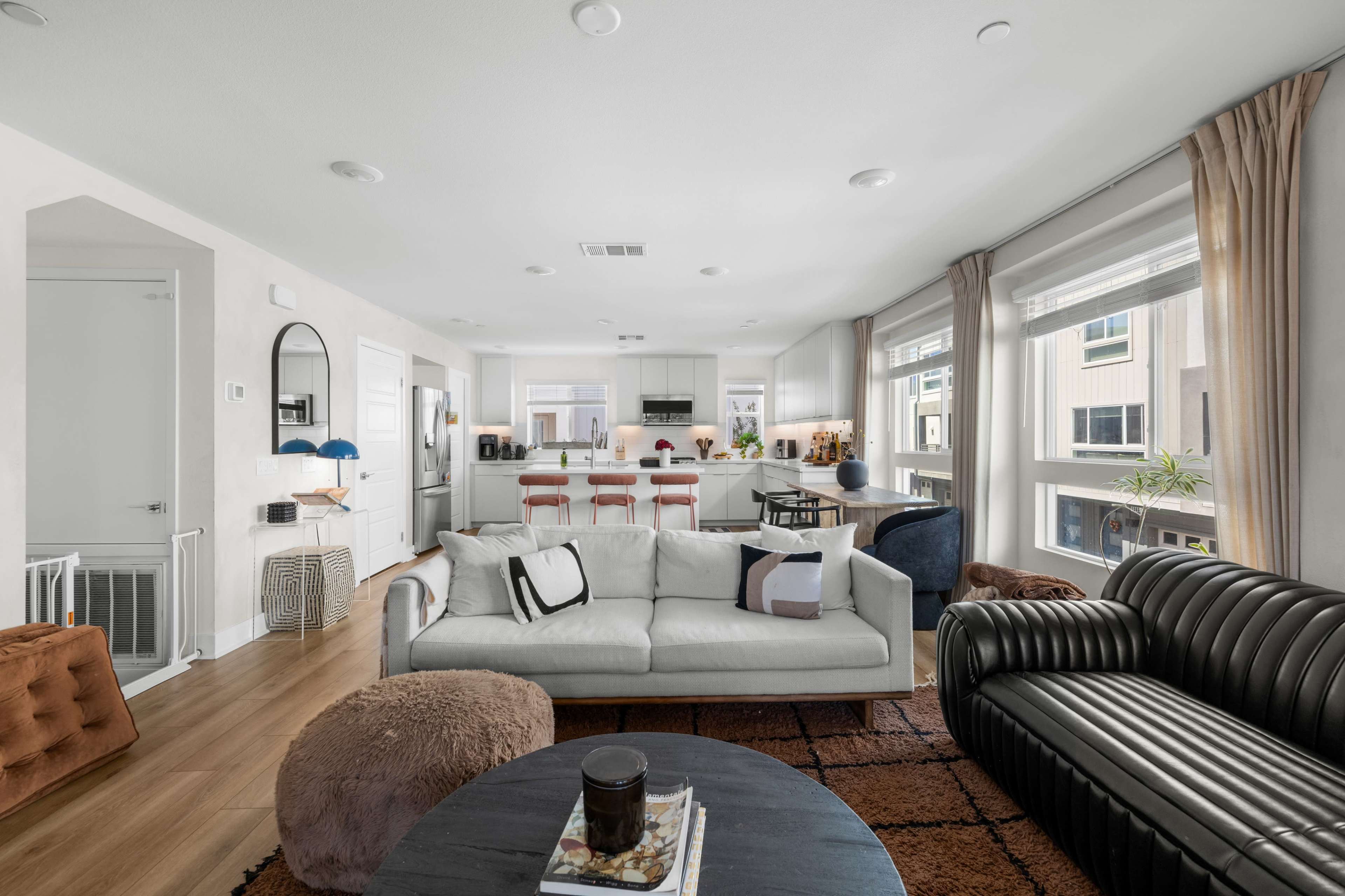 The image shows a modern living room area connected to a kitchen, featuring a gray couch, a black leather sofa, and a circular coffee table.