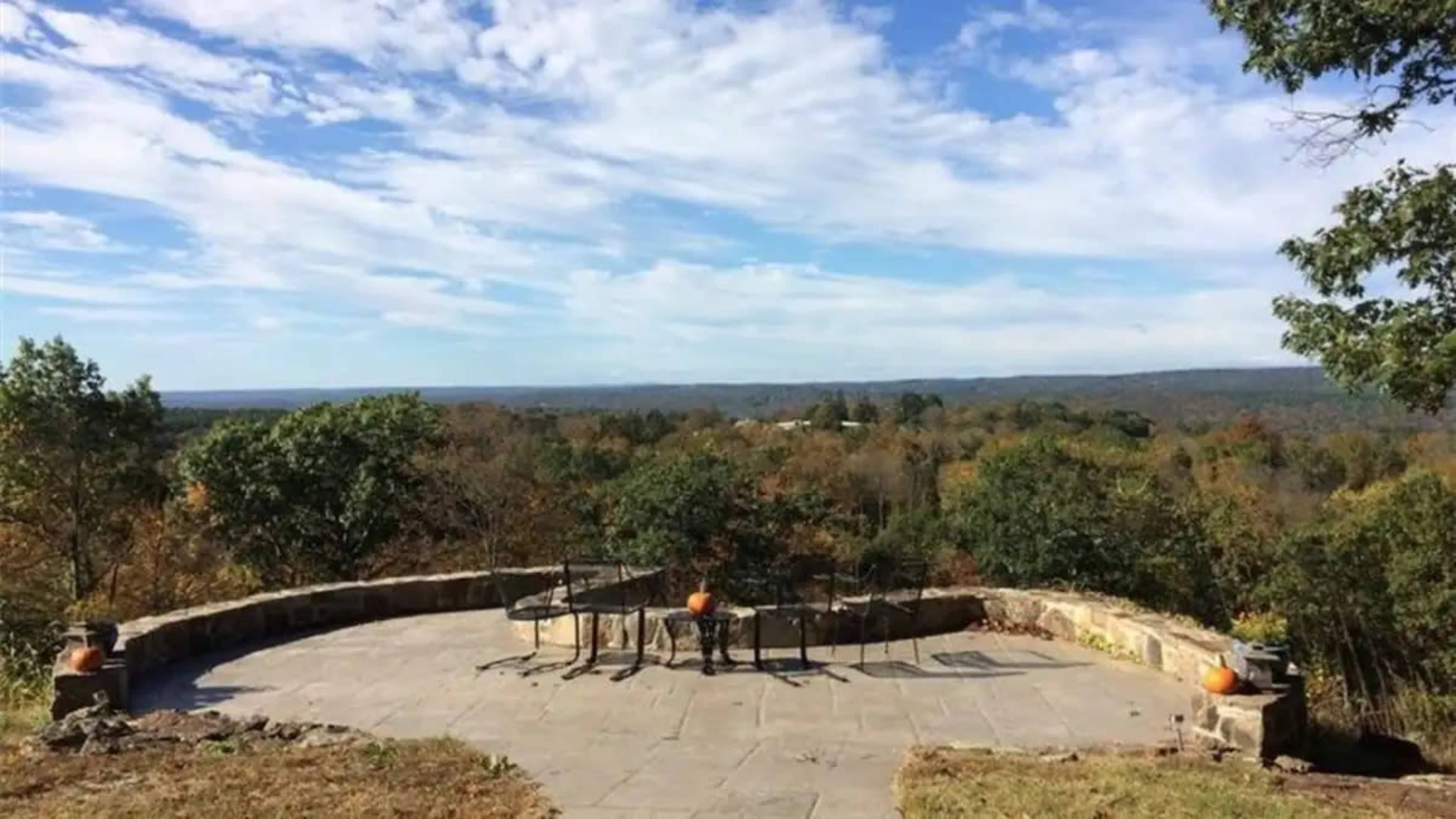 A stone patio with a table and chairs overlooks a wooded landscape under a cloudy sky.