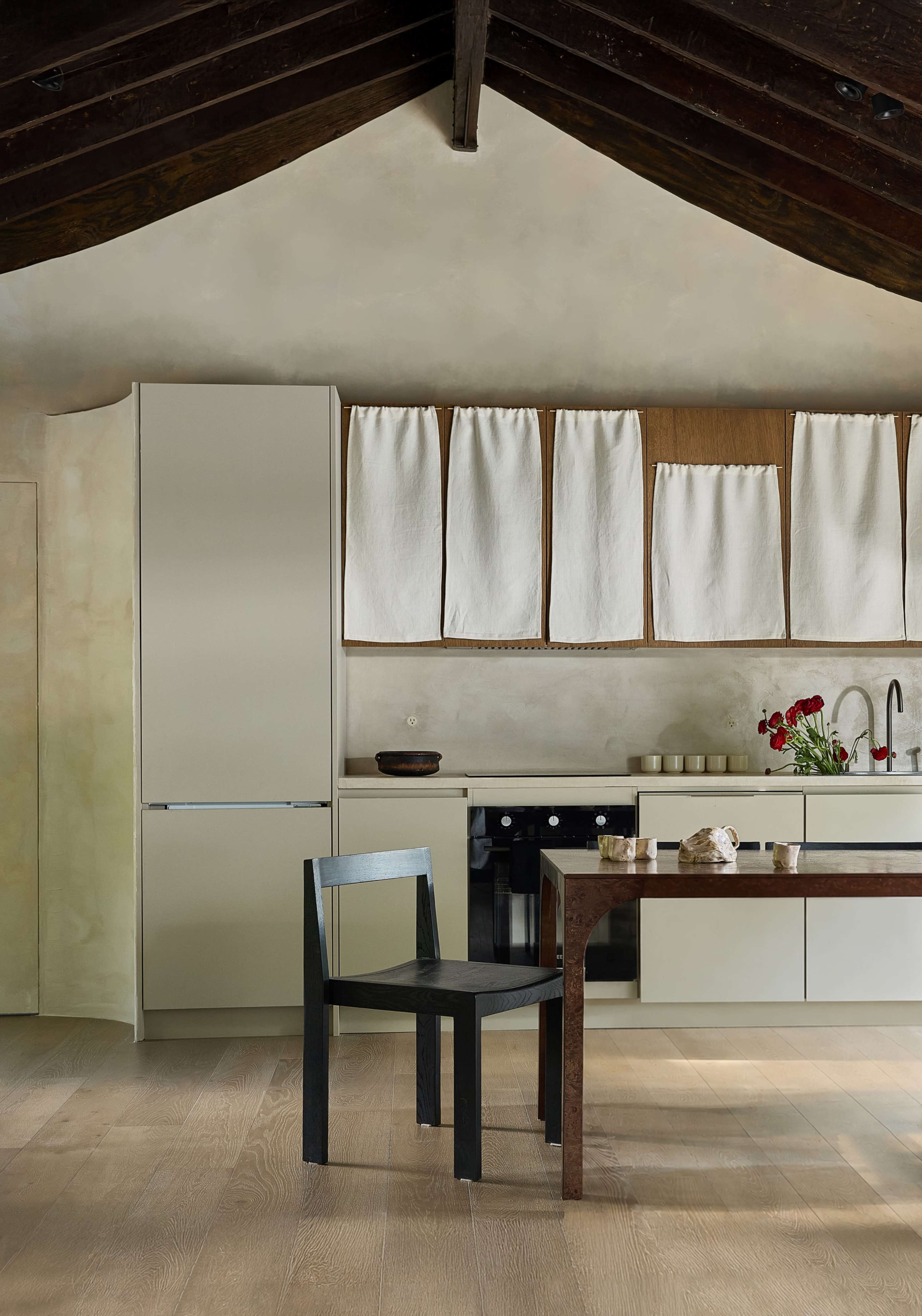The image shows a modern kitchen with a minimalist design, featuring a light-colored cabinetry, a wooden dining table, and a single black chair.