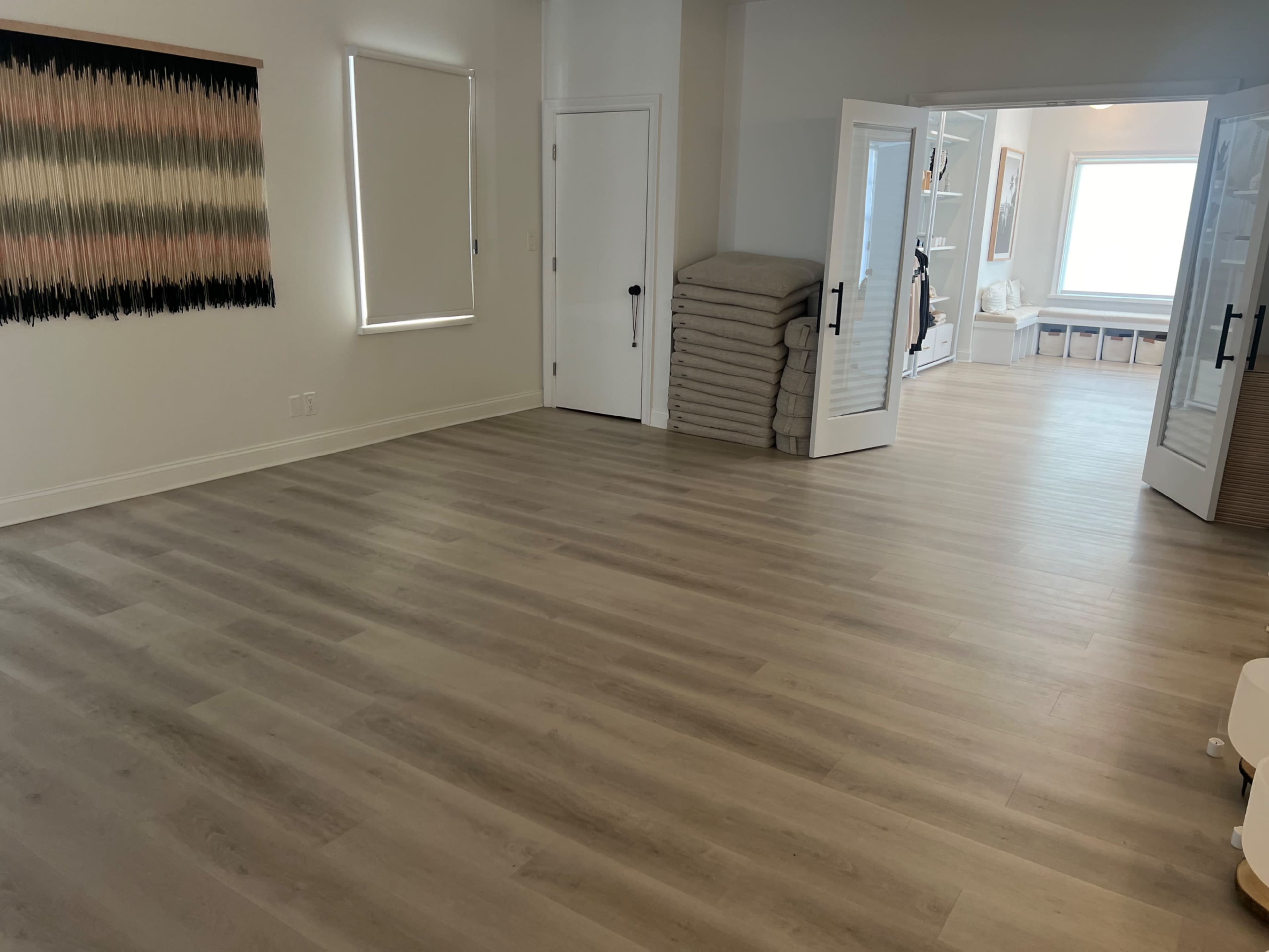 The room features a light wood floor with neatly stacked blankets against one wall and a doorway leading to a bright, furnished space.