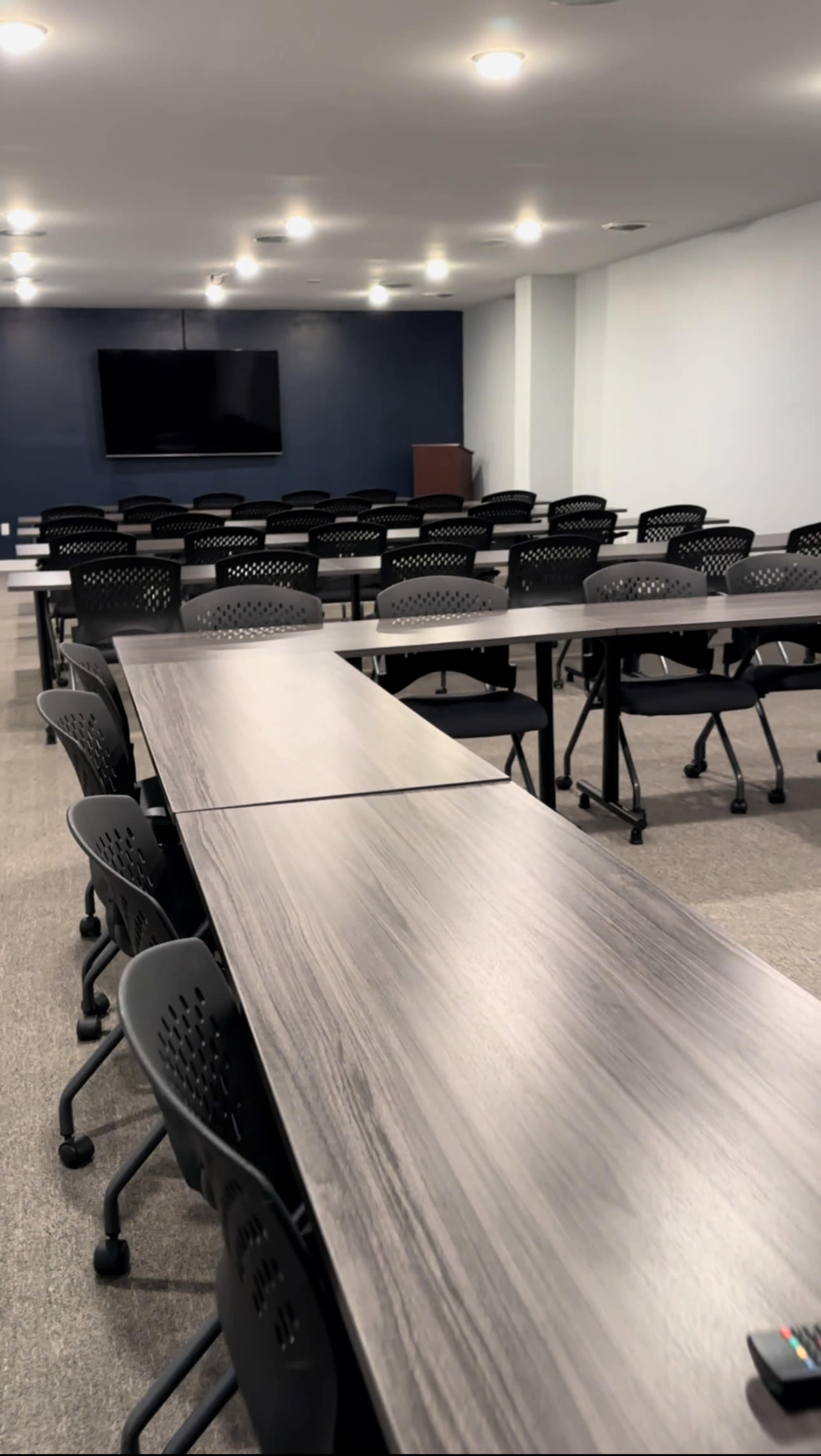 A modern meeting room features multiple tables arranged in a U-shape, accompanied by rolling chairs and a large screen on the wall.