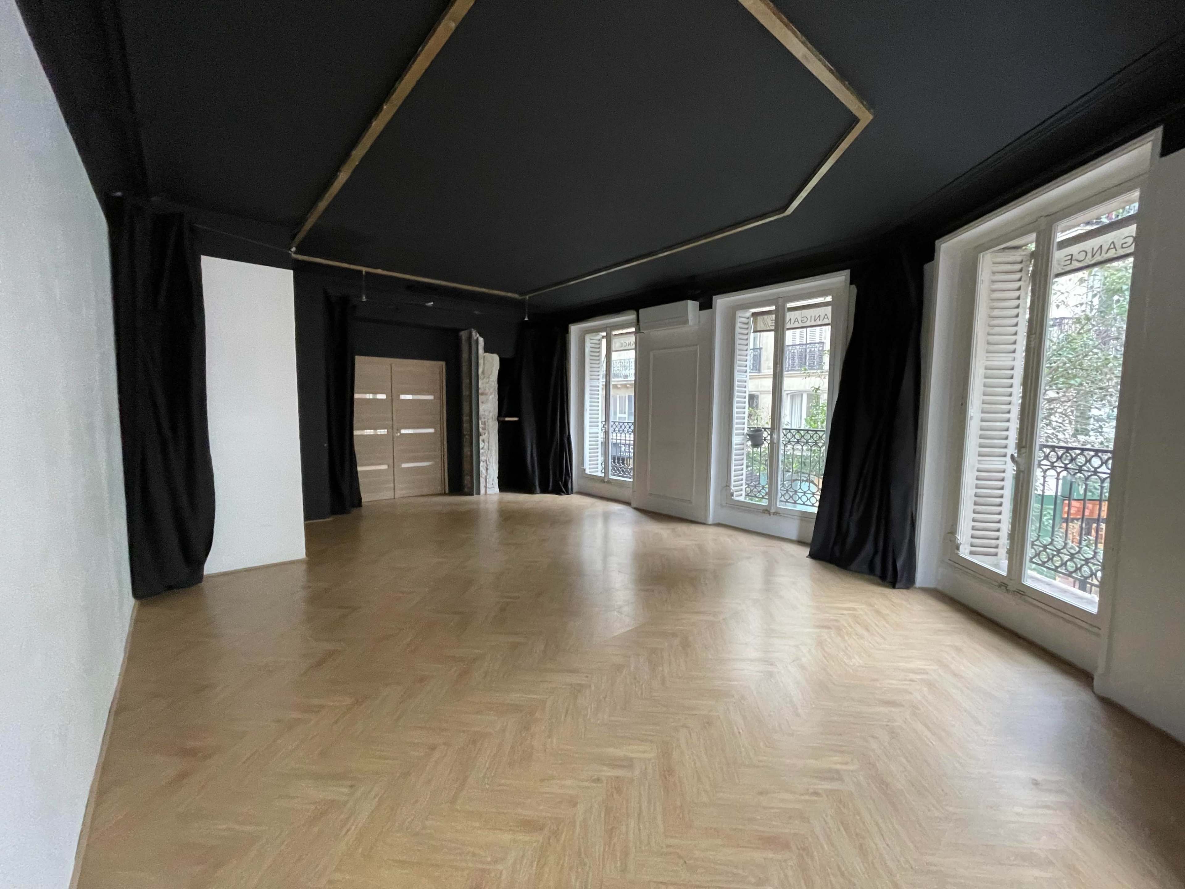 An empty room with a dark ceiling, herringbone wooden flooring, and large windows covered with black curtains.