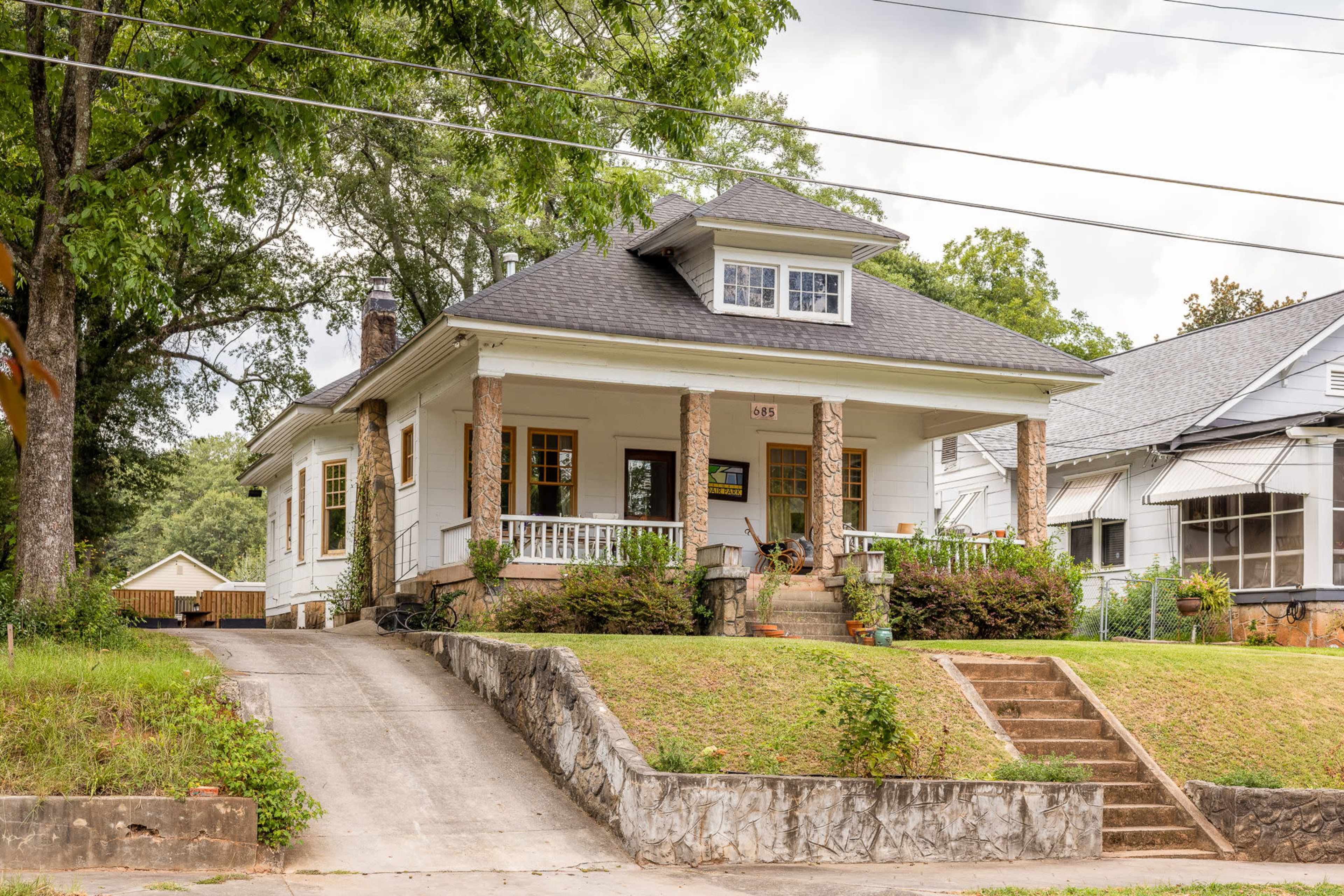 A single-story house with a gabled roof features a front porch supported by stone columns and is set on a grassy yard with a gravel driveway.