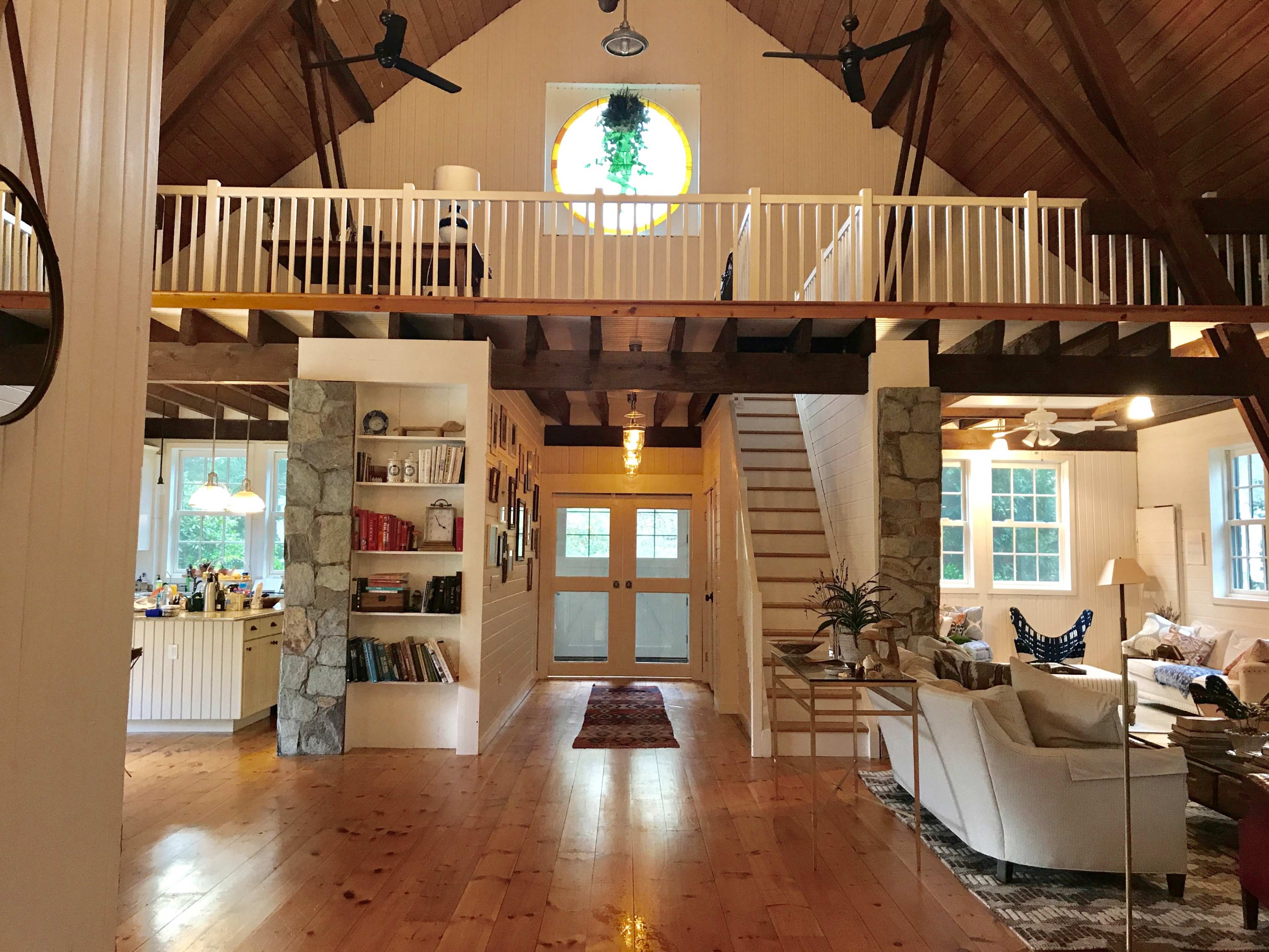 A spacious living area with a high wooden ceiling, featuring a staircase, large windows, and a balcony overlooking the main floor.