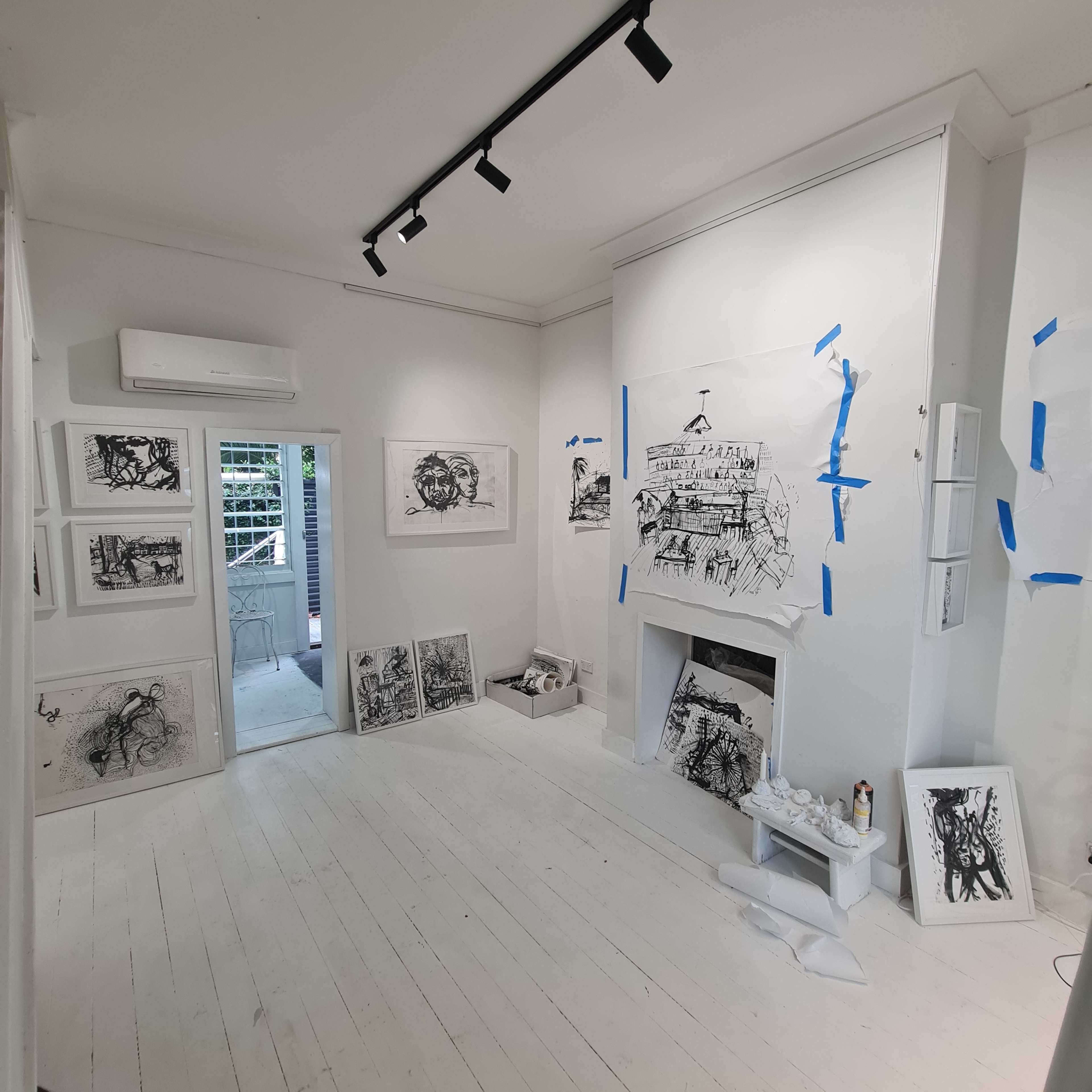 An art studio featuring various black and white drawings on the walls and floor, with blue tape securing some of the artworks.