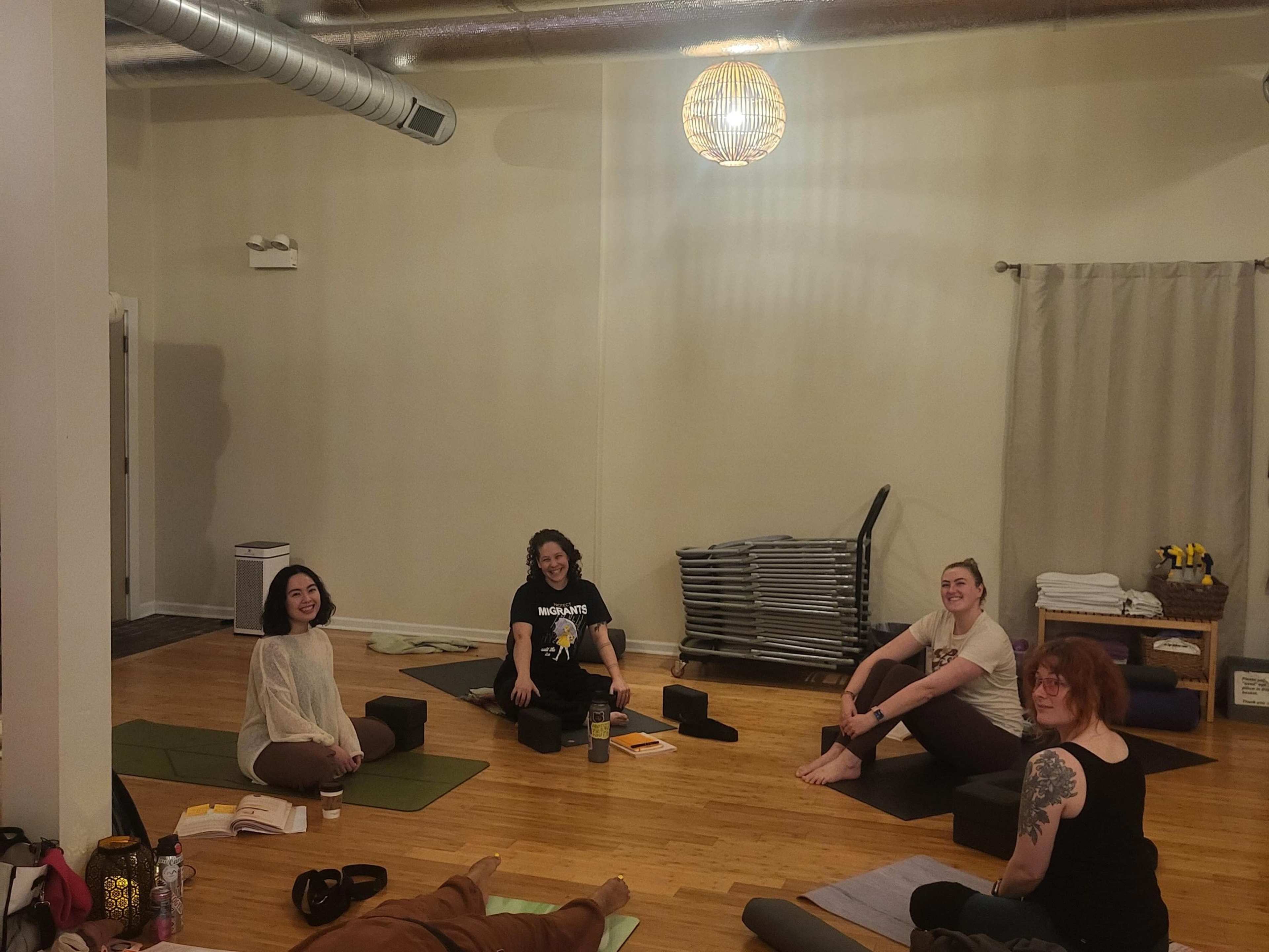 A group of five individuals is seated on yoga mats in a bright studio, with equipment stacked in the background.