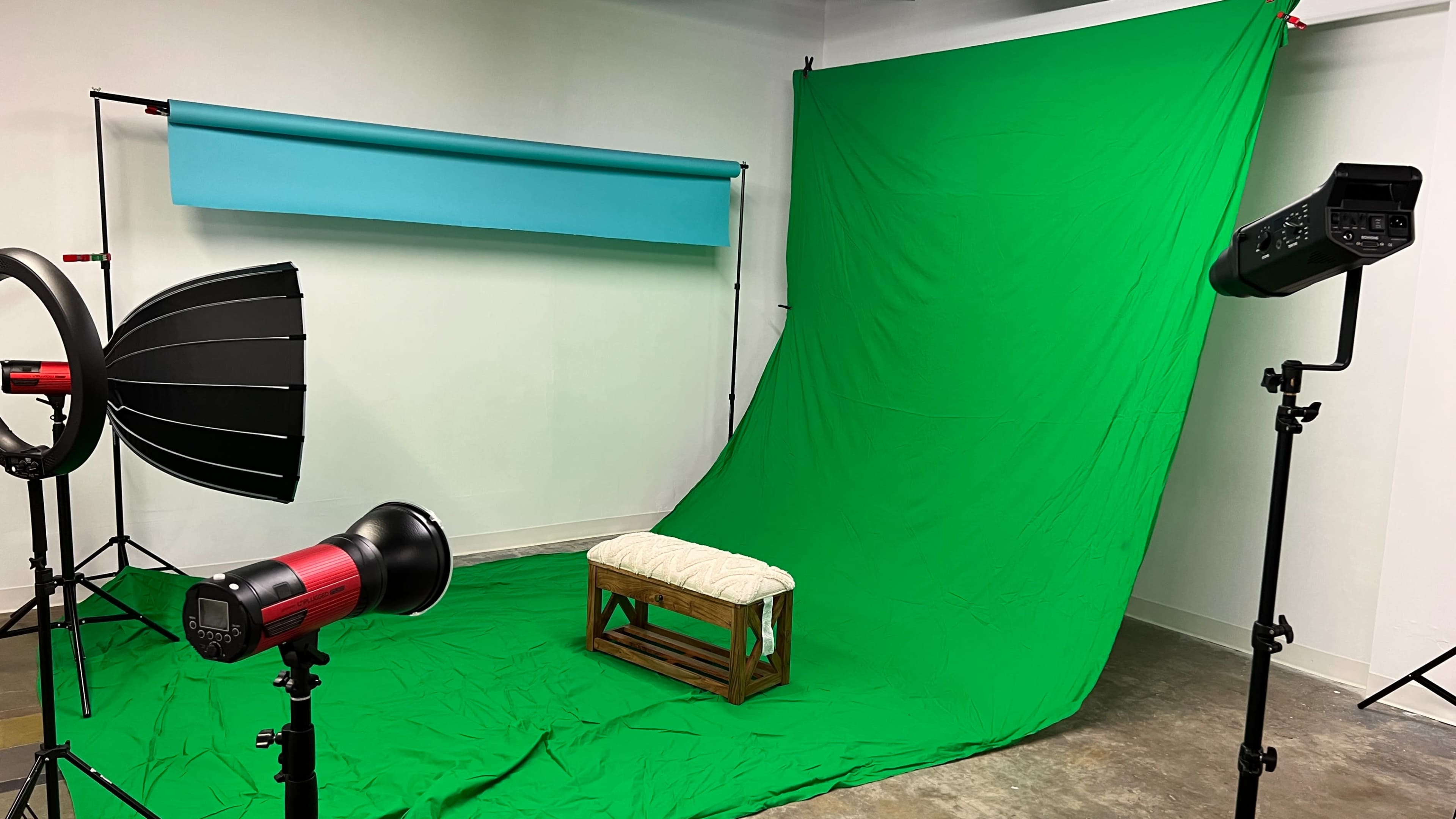 A photography studio with a green backdrop, a blue overhead screen, and lighting equipment positioned around a wooden bench.