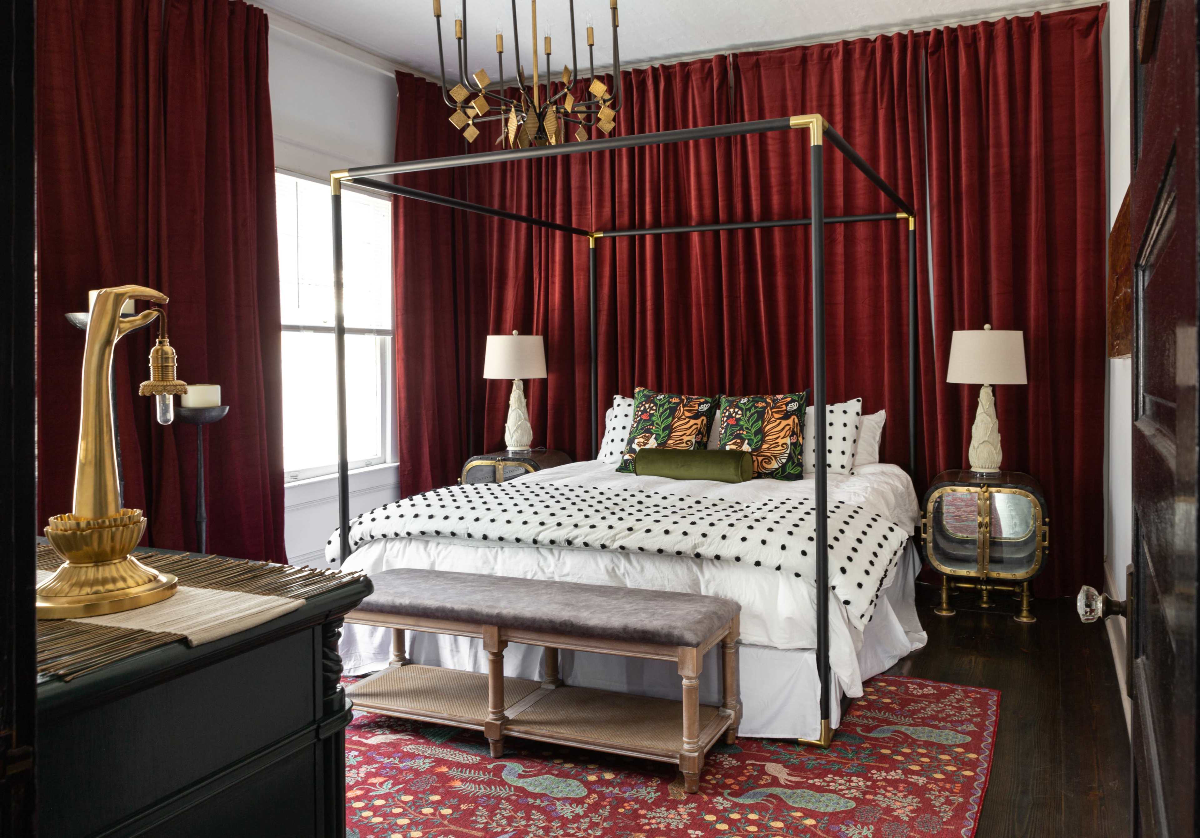 A bedroom featuring a four-poster bed with a bold black frame, polka dot bedding, and rich red curtains, complemented by vintage lamps and a patterned area rug.