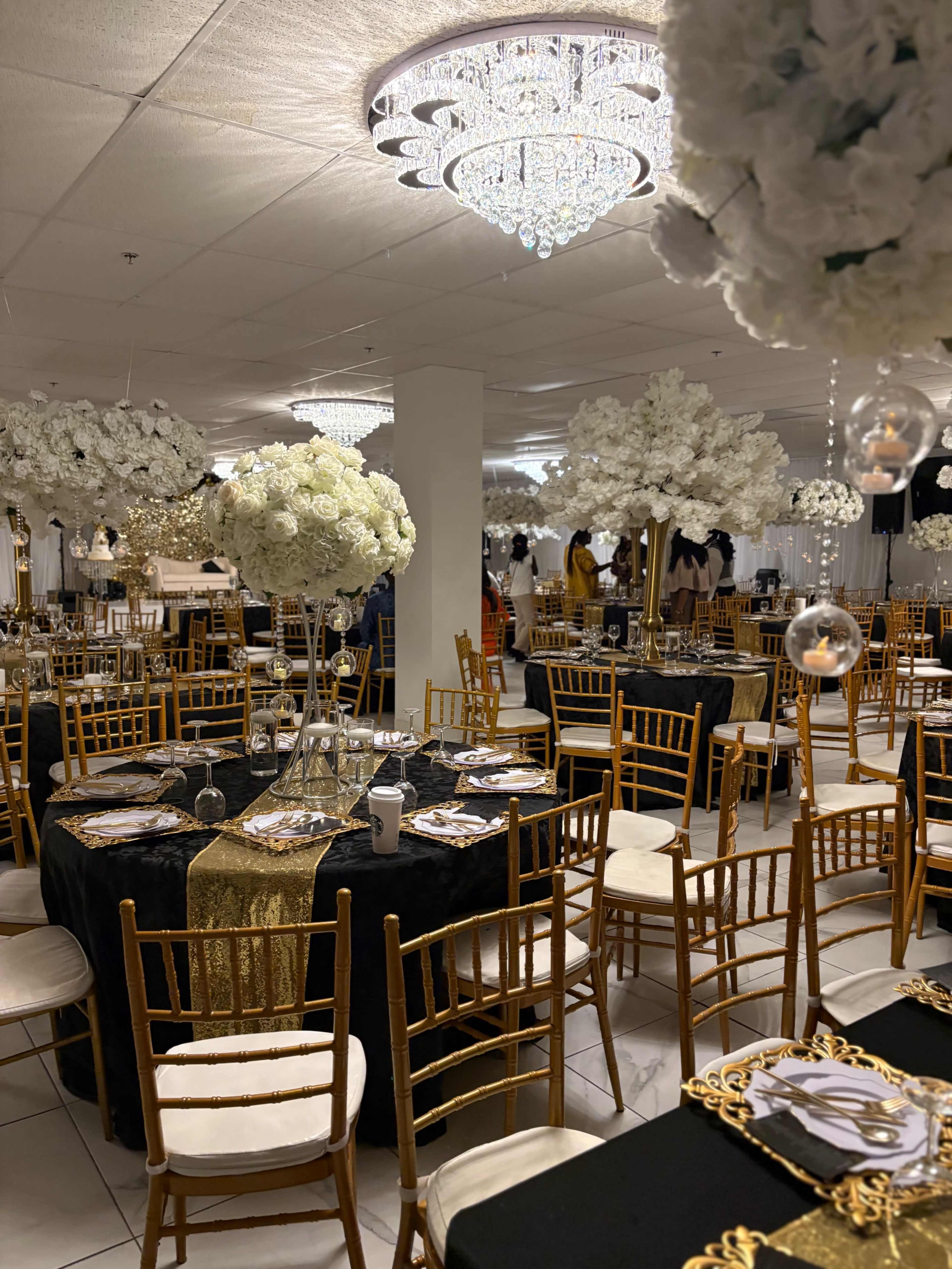 The image shows an elegantly decorated banquet hall with round tables draped in black and gold, adorned with floral centerpieces and crystal chandeliers overhead.