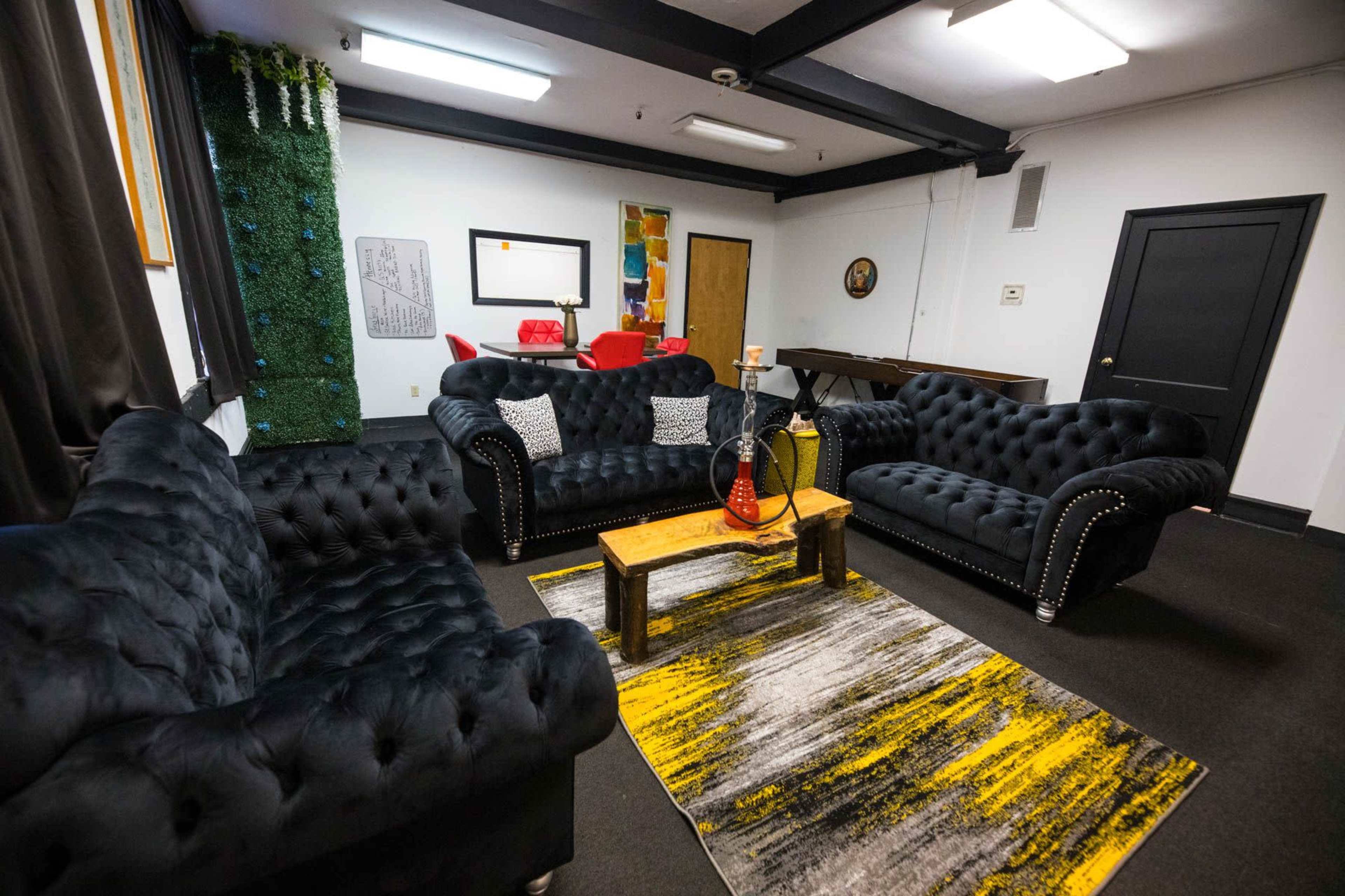 The room features black tufted sofas arranged around a wooden coffee table on a patterned rug, with a mirror and colorful decorations on the walls.