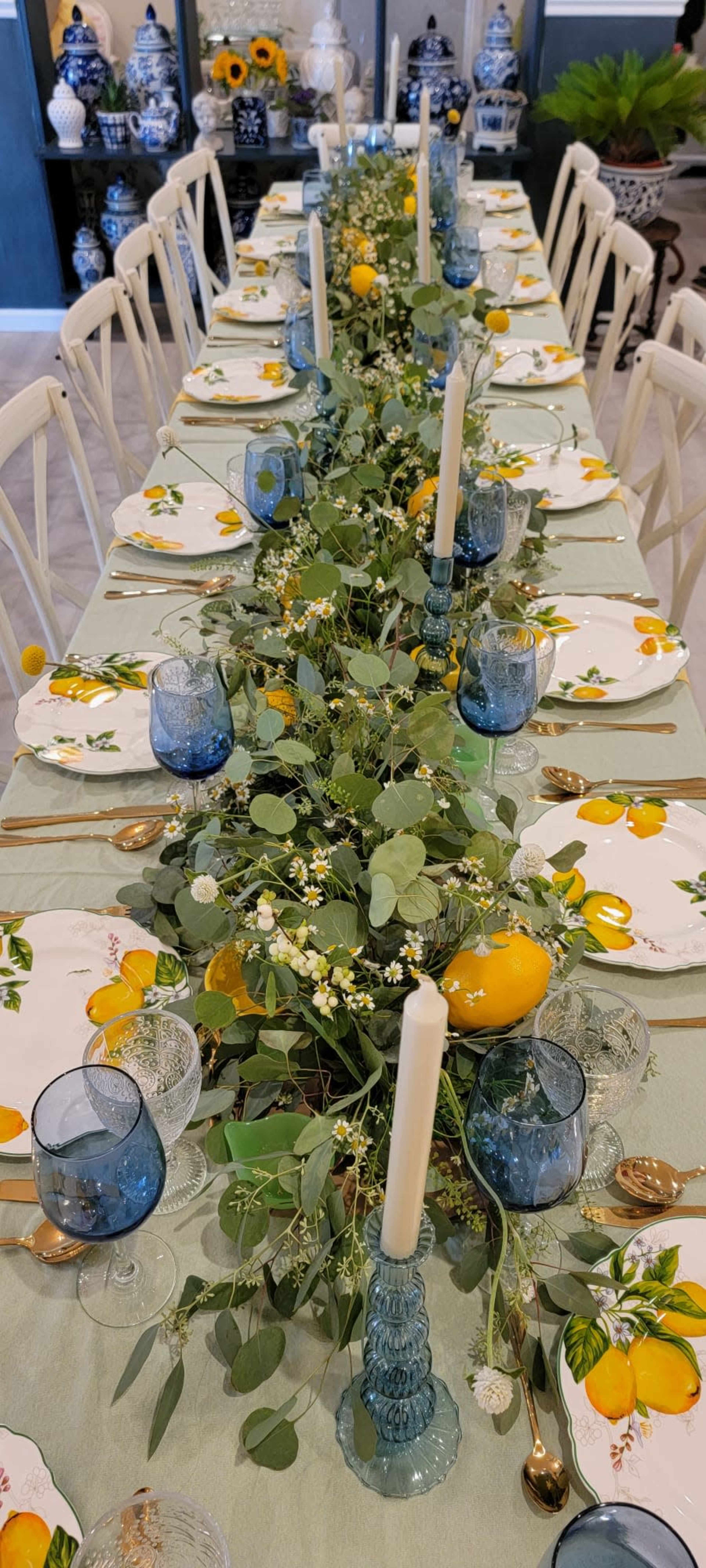 A long dining table is elegantly arranged with lemon-themed tableware, greenery, and blue glasses, centered around a vibrant floral centerpiece.