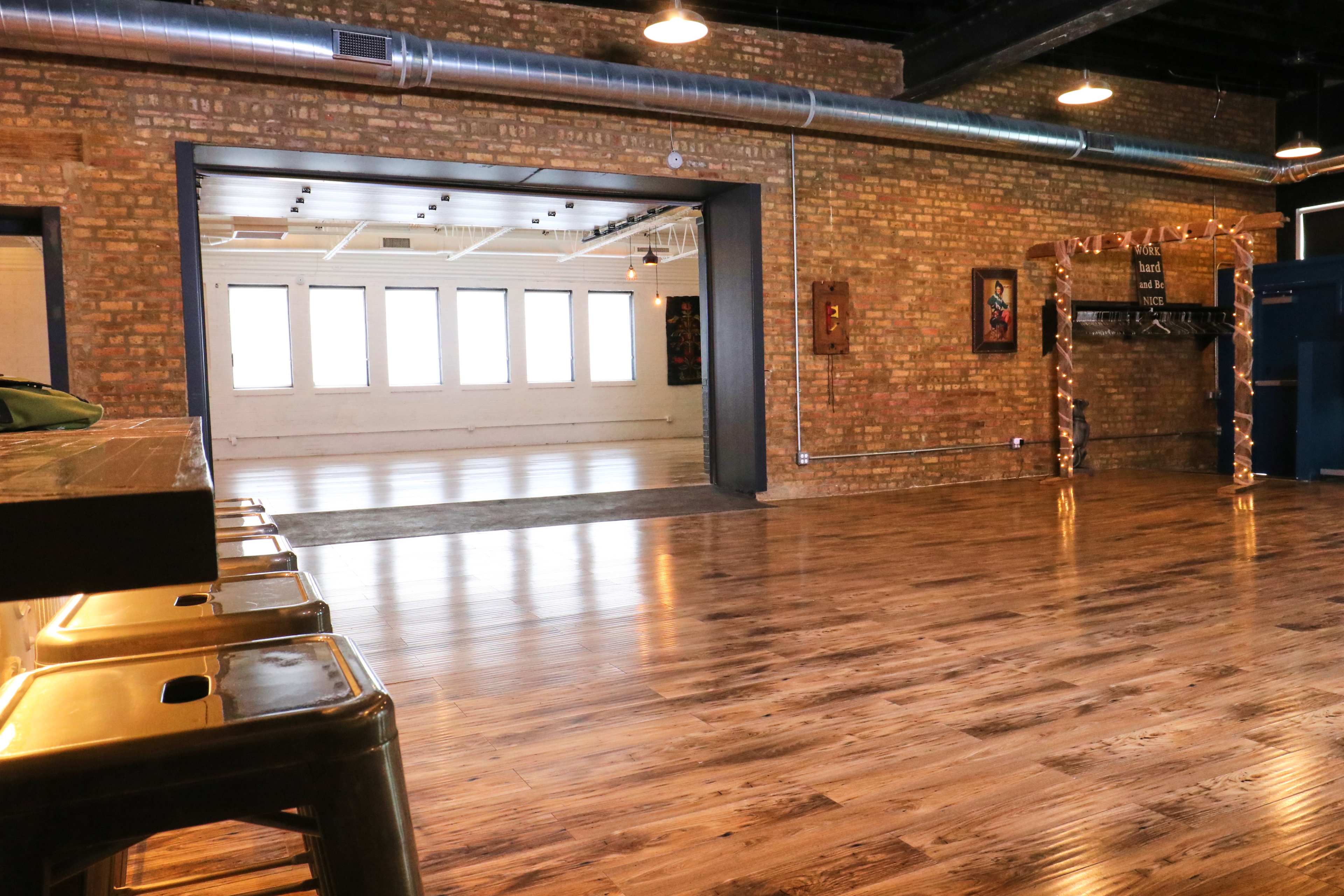 The image shows a spacious room with polished wooden floors and large windows, separated by an open doorway, with exposed brick walls and metal bar stools.