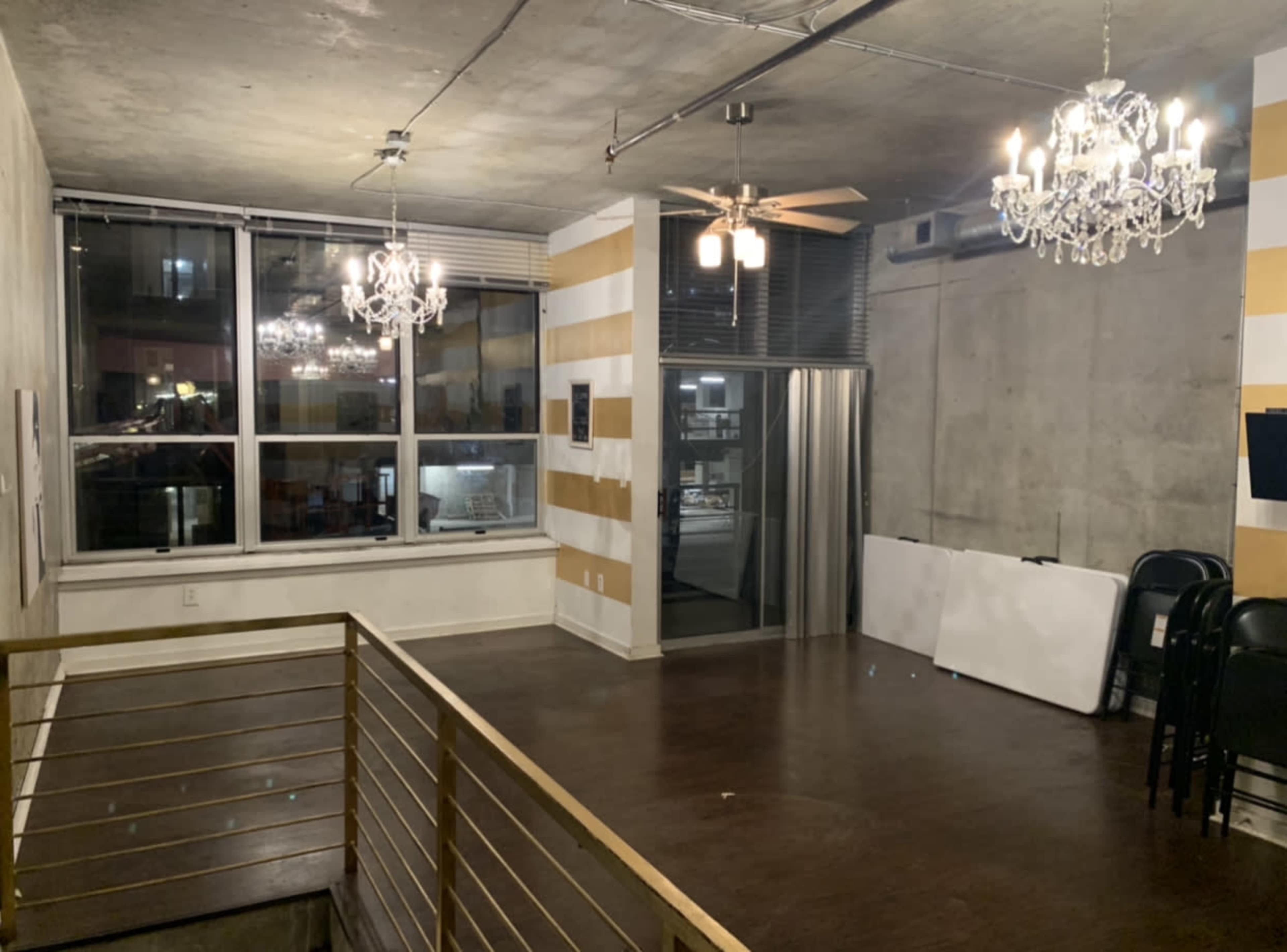 A spacious room with concrete walls, wooden flooring, two chandelier light fixtures, and a large window overlooking an outdoor area.
