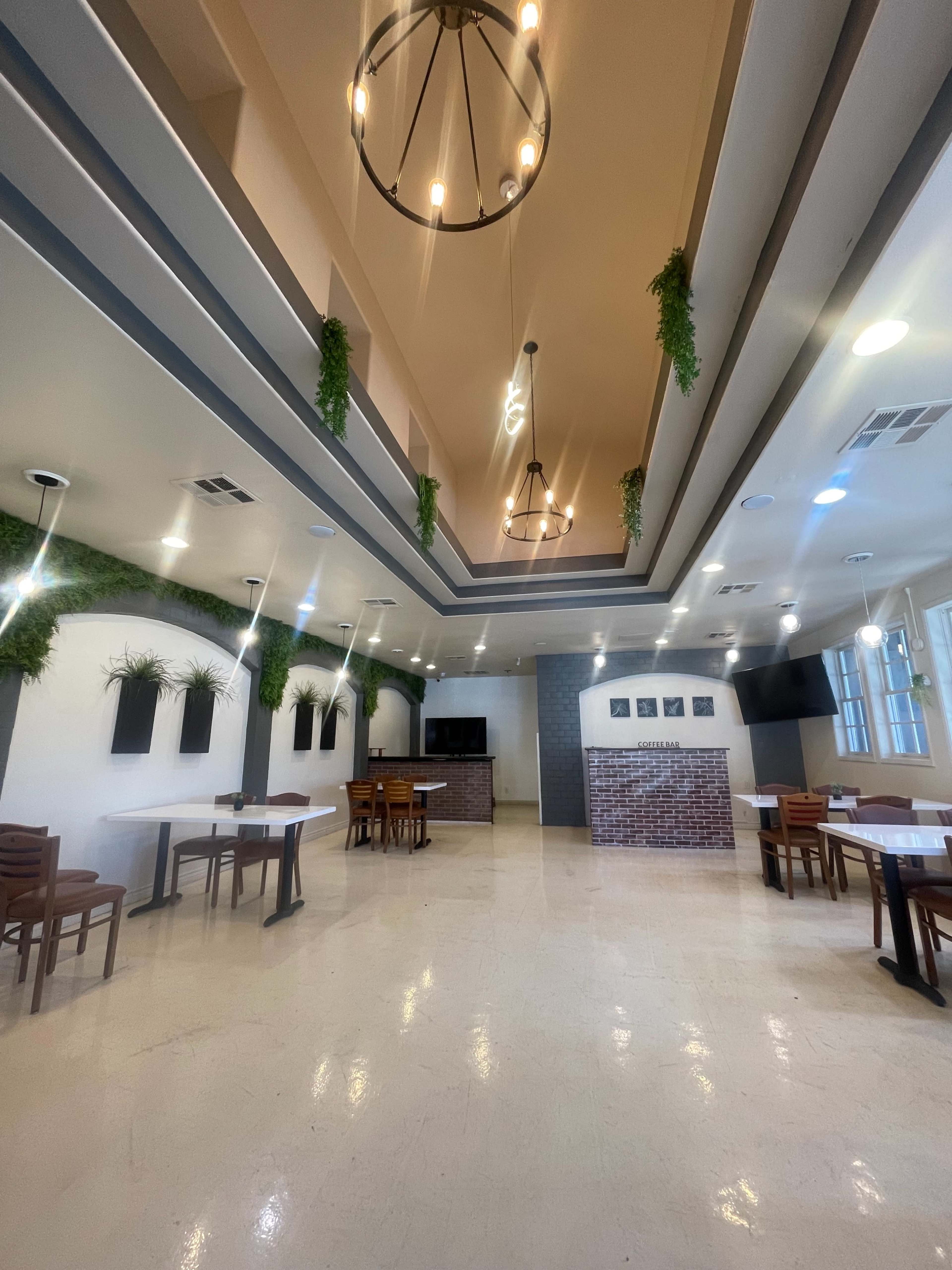 A spacious restaurant interior featuring a high ceiling, modern lighting fixtures, and plants on the walls, with several tables and chairs arranged throughout.