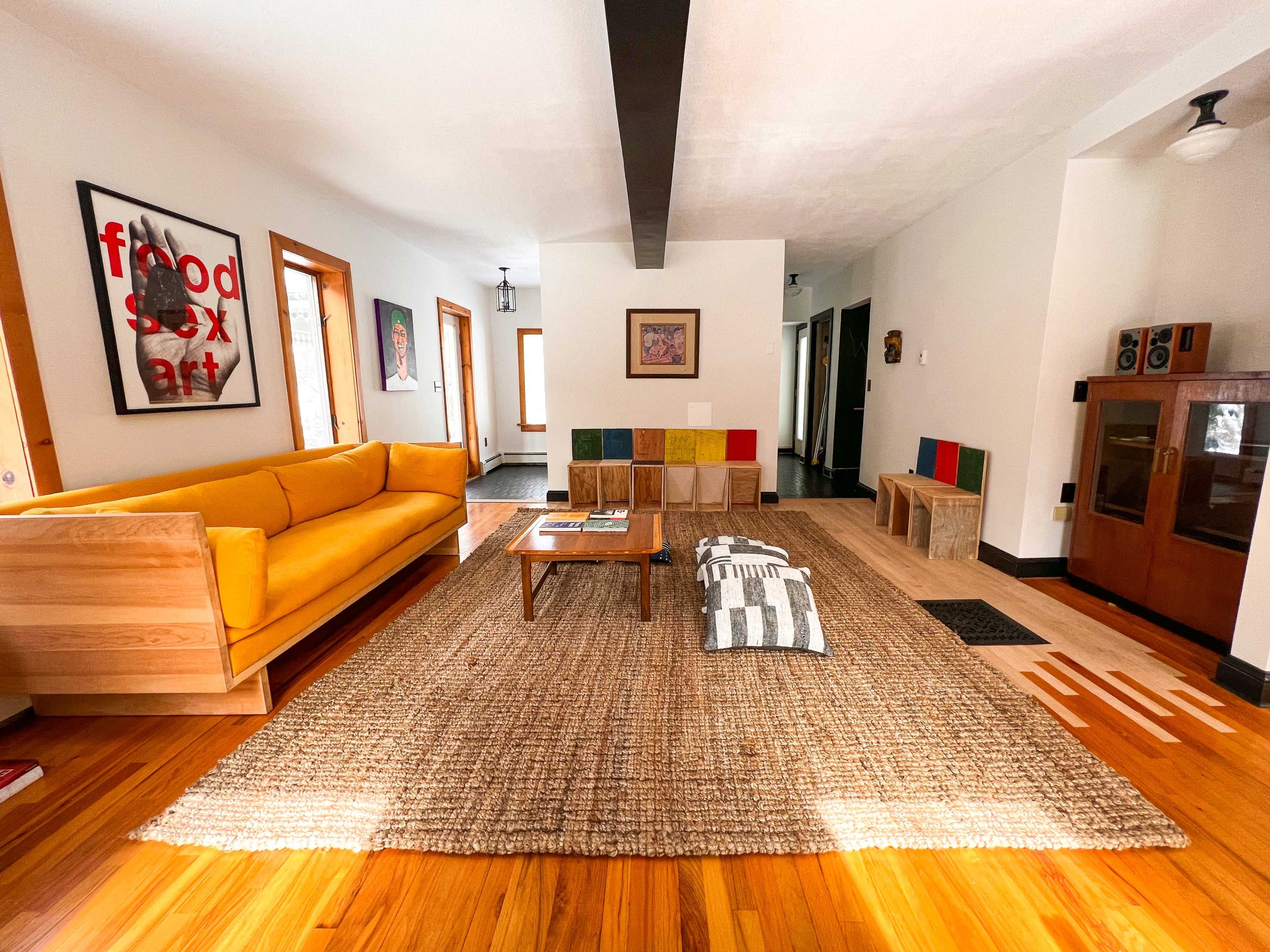 The scene shows a bright living room featuring a yellow sofa, a large area rug, a wooden coffee table, and colorful wall decorations.