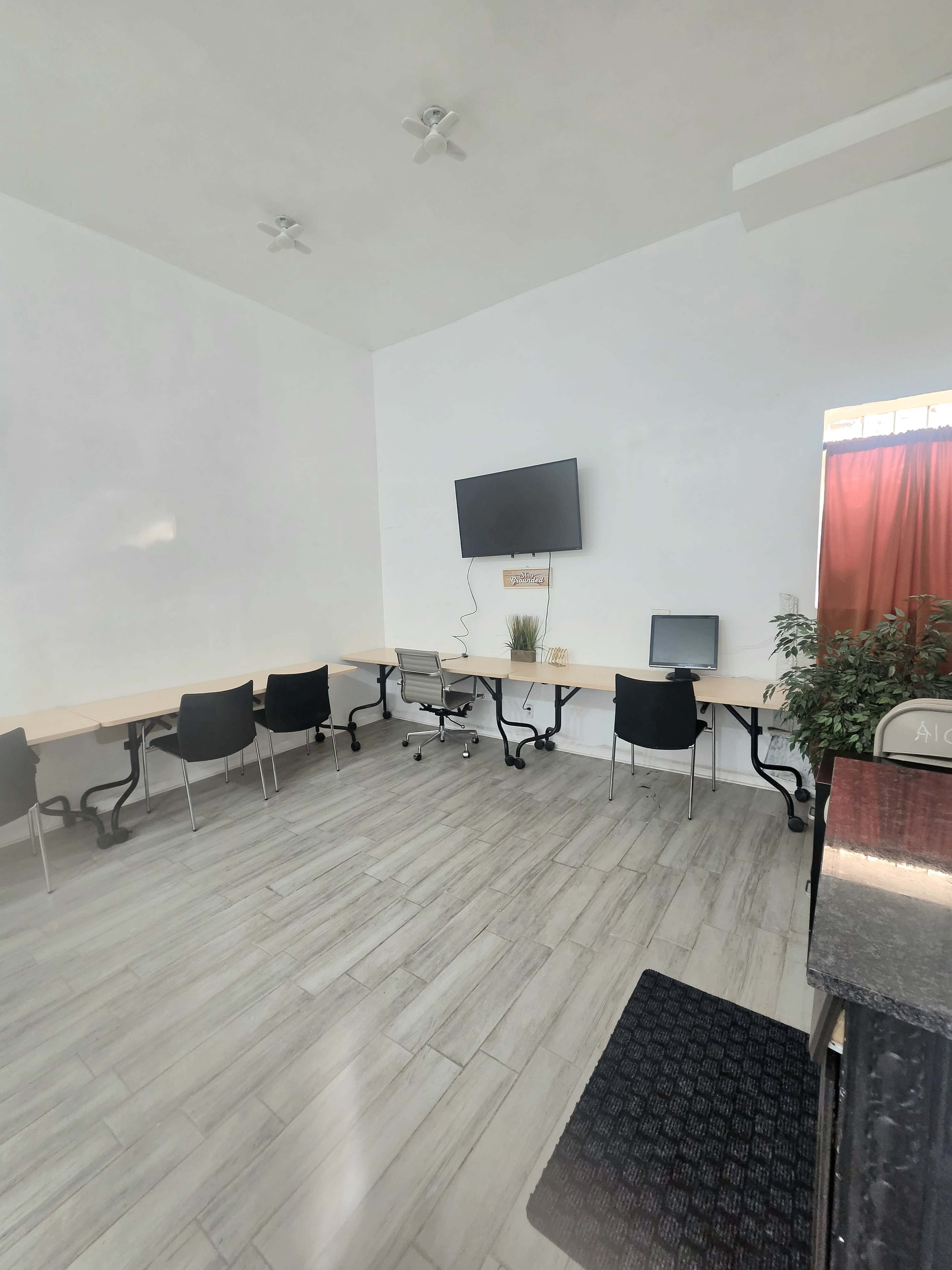 The image shows a bright, spacious room with several tables and chairs arranged along the walls, a television mounted on one wall, and a potted plant near the window.