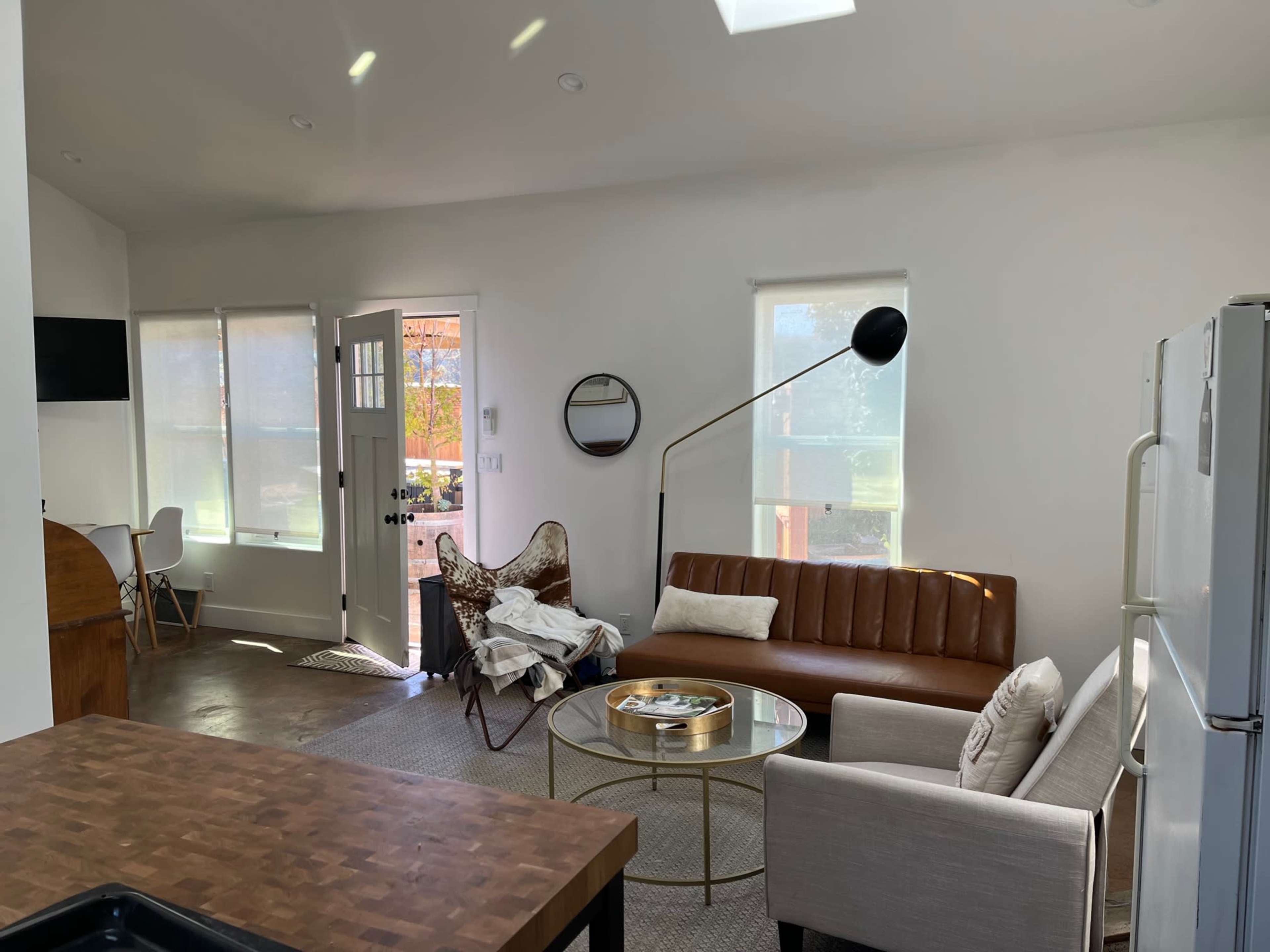 A modern living room features a brown leather sofa, a beige armchair, and a coffee table, with large windows allowing natural light to enter.