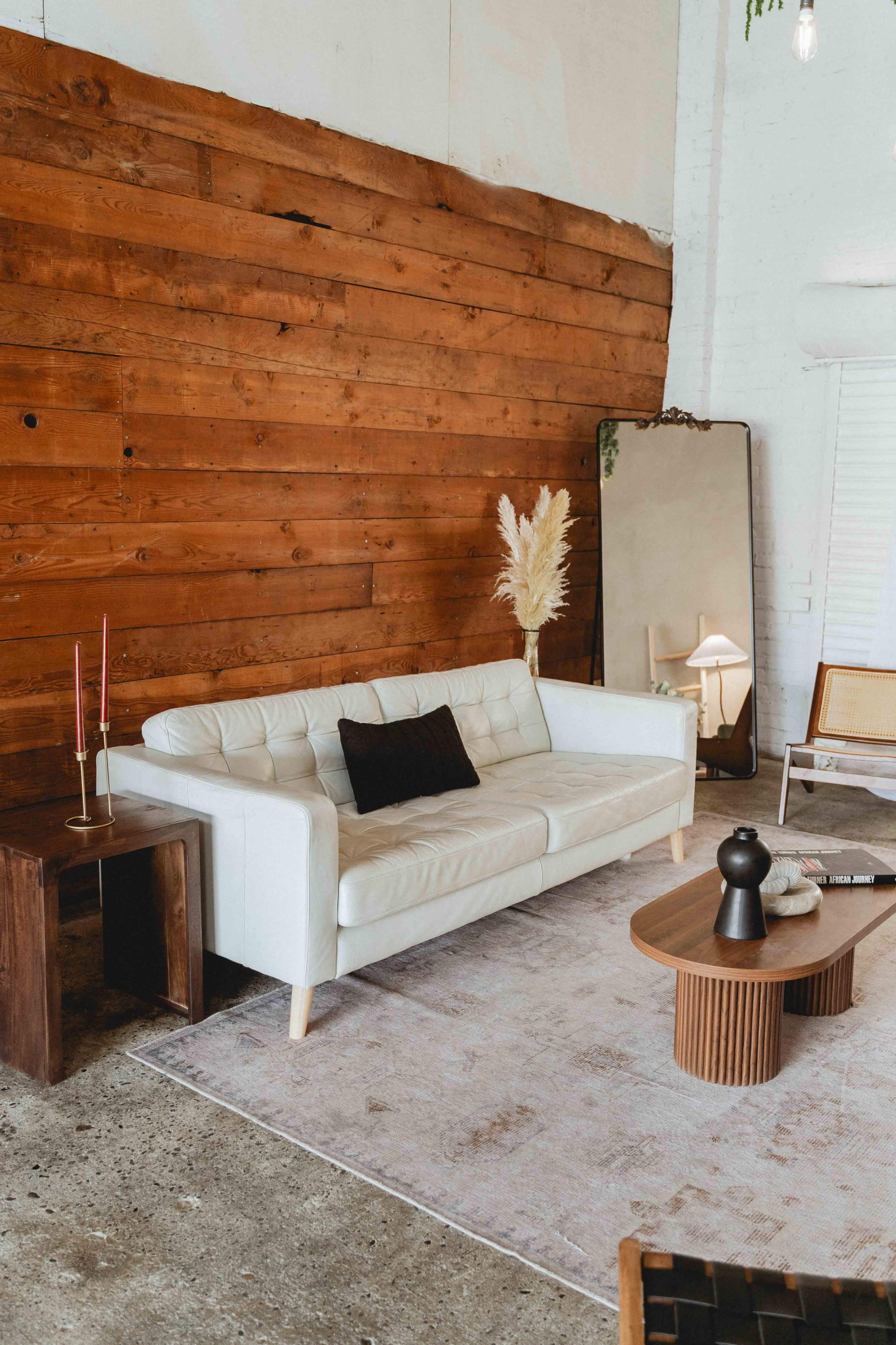 The image shows a modern living room with a white leather sofa, a wooden wall, a large mirror, a small table with decorative items, and a rug on a concrete floor.
