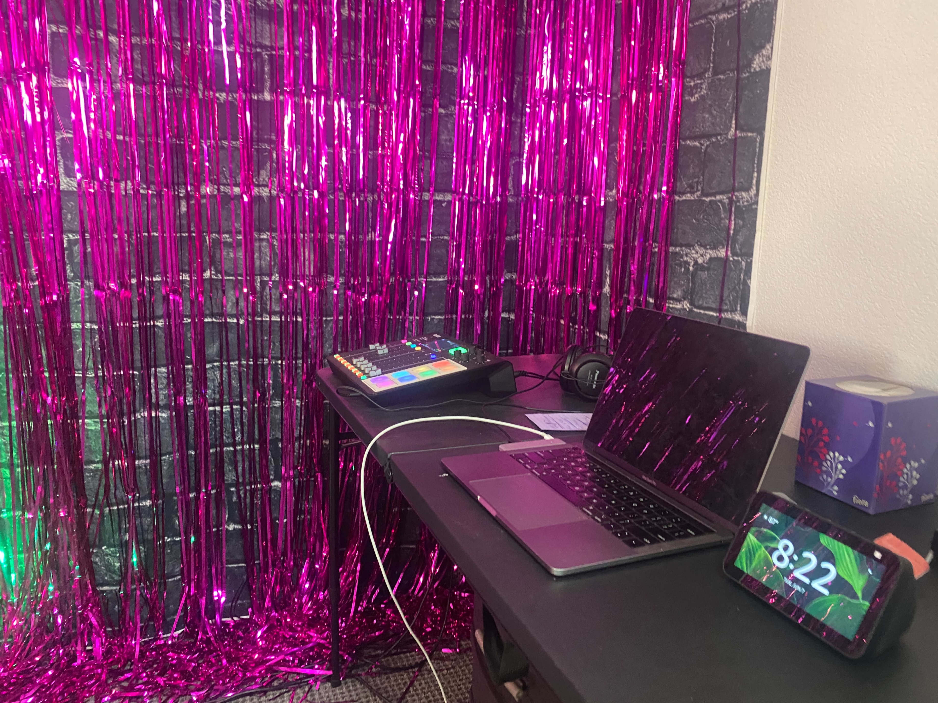A desk with a laptop, a sound control device, and a smartphone is set against a shiny pink foiled backdrop.