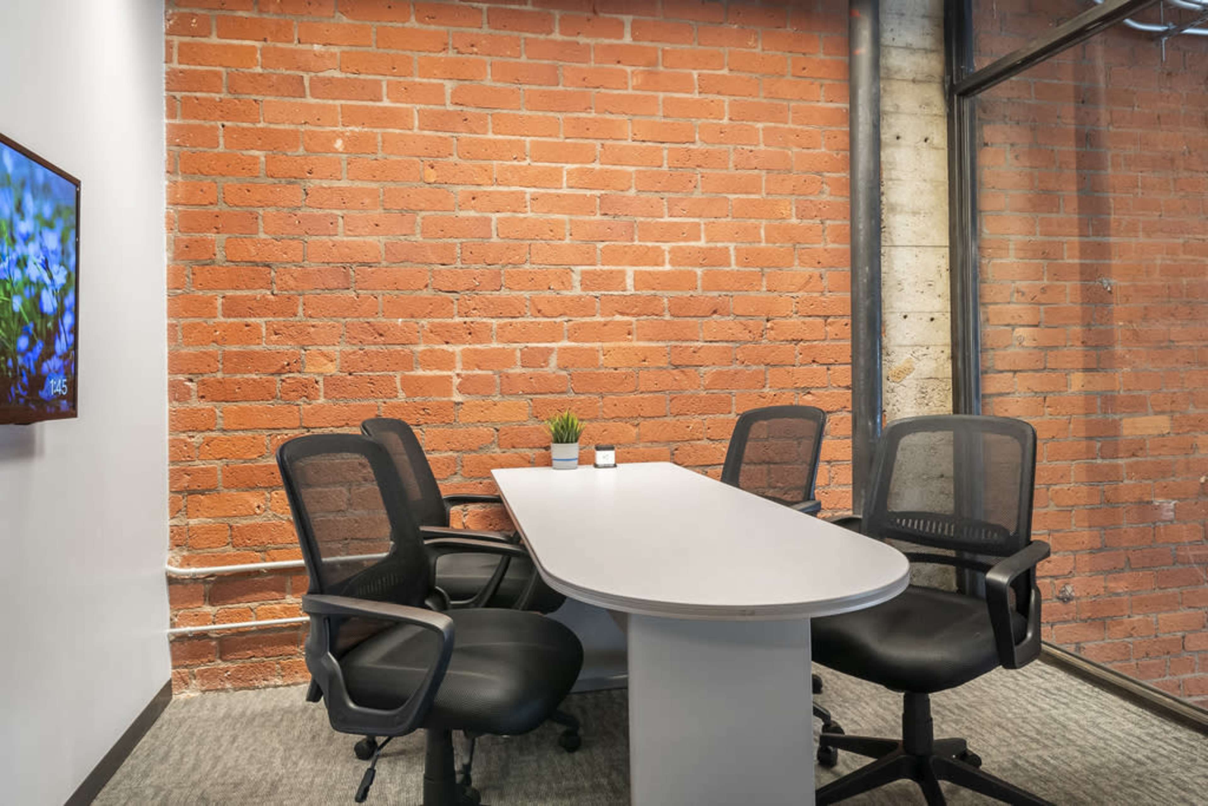 A modern meeting room with a round white table and three black office chairs, set against a backdrop of exposed brick walls.