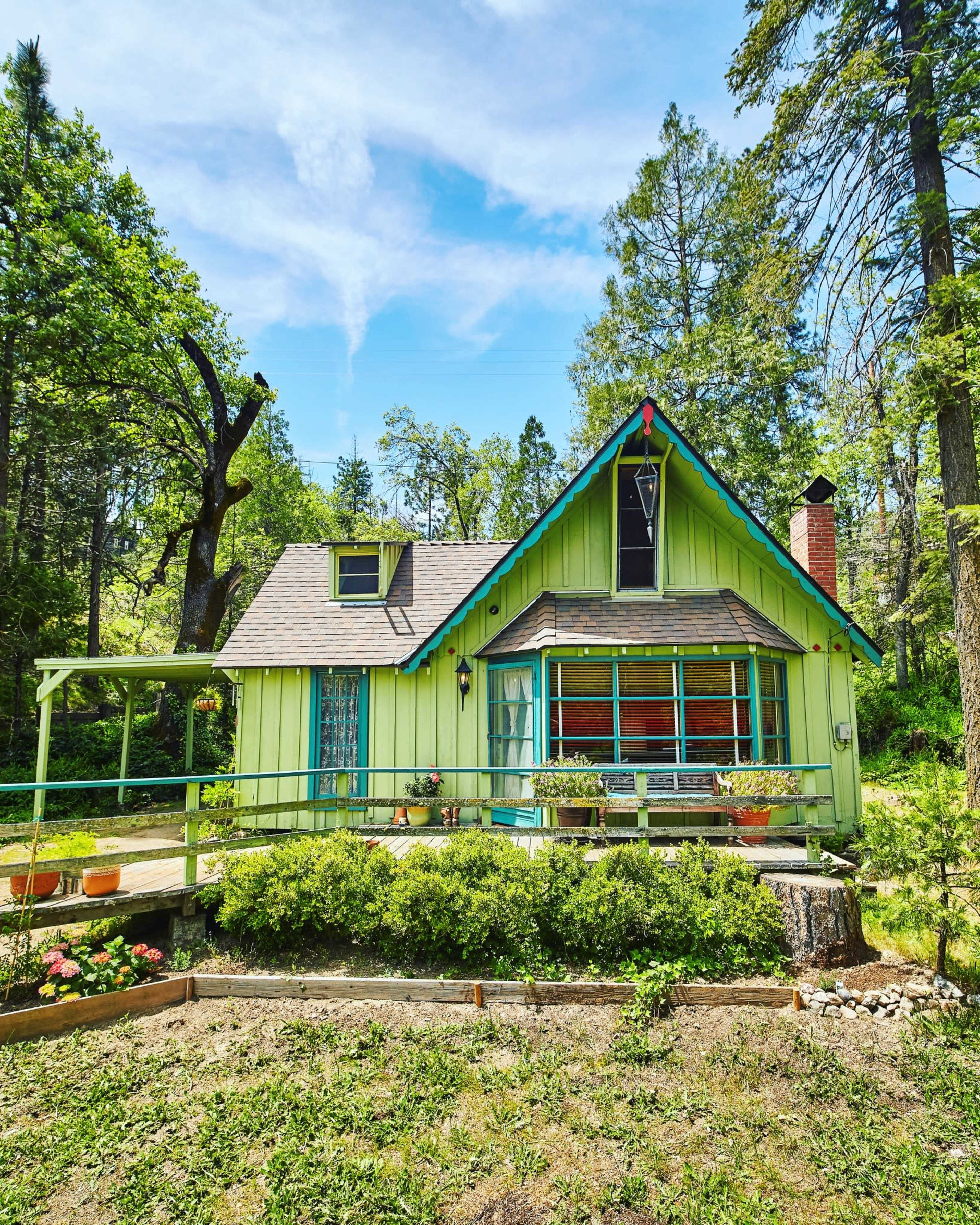 A green, A-frame cottage with large windows sits surrounded by trees and garden beds in a rural setting.