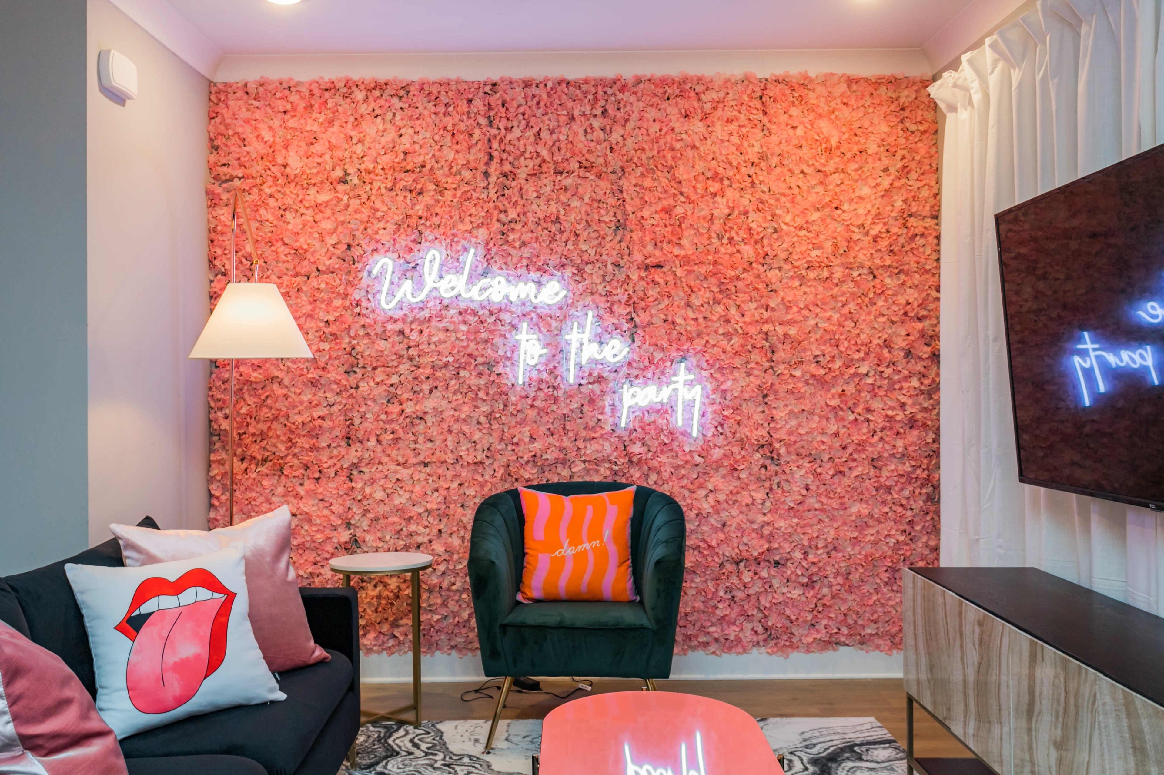 The scene features a modern living room with a wall covered in pink floral decorations and neon signs that read "Welcome to the party."