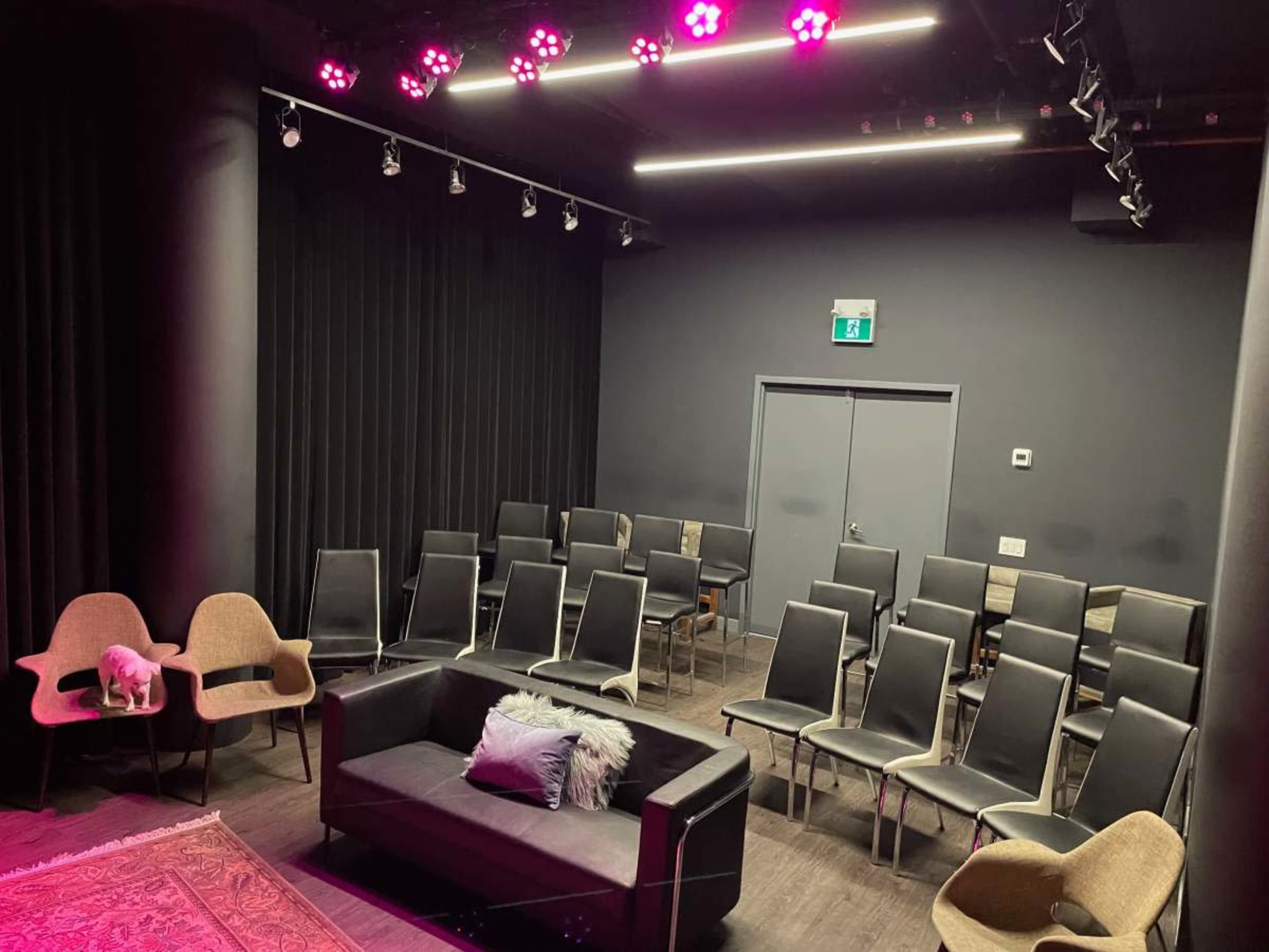 The image shows a dimly lit performance space with rows of black chairs facing a small stage area that features a sofa and two light-colored armchairs.