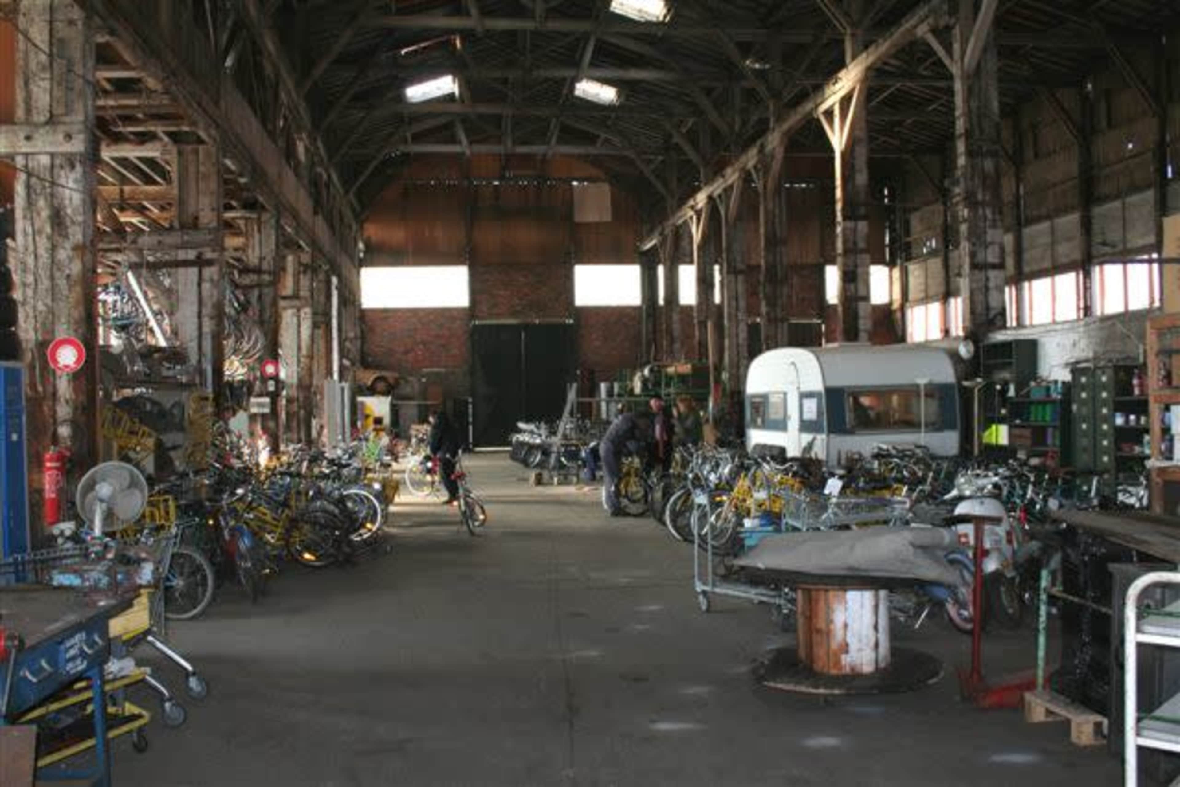 The image shows a large, industrial warehouse filled with rows of bicycles and various tools, with exposed wooden beams and a few people working in the space.