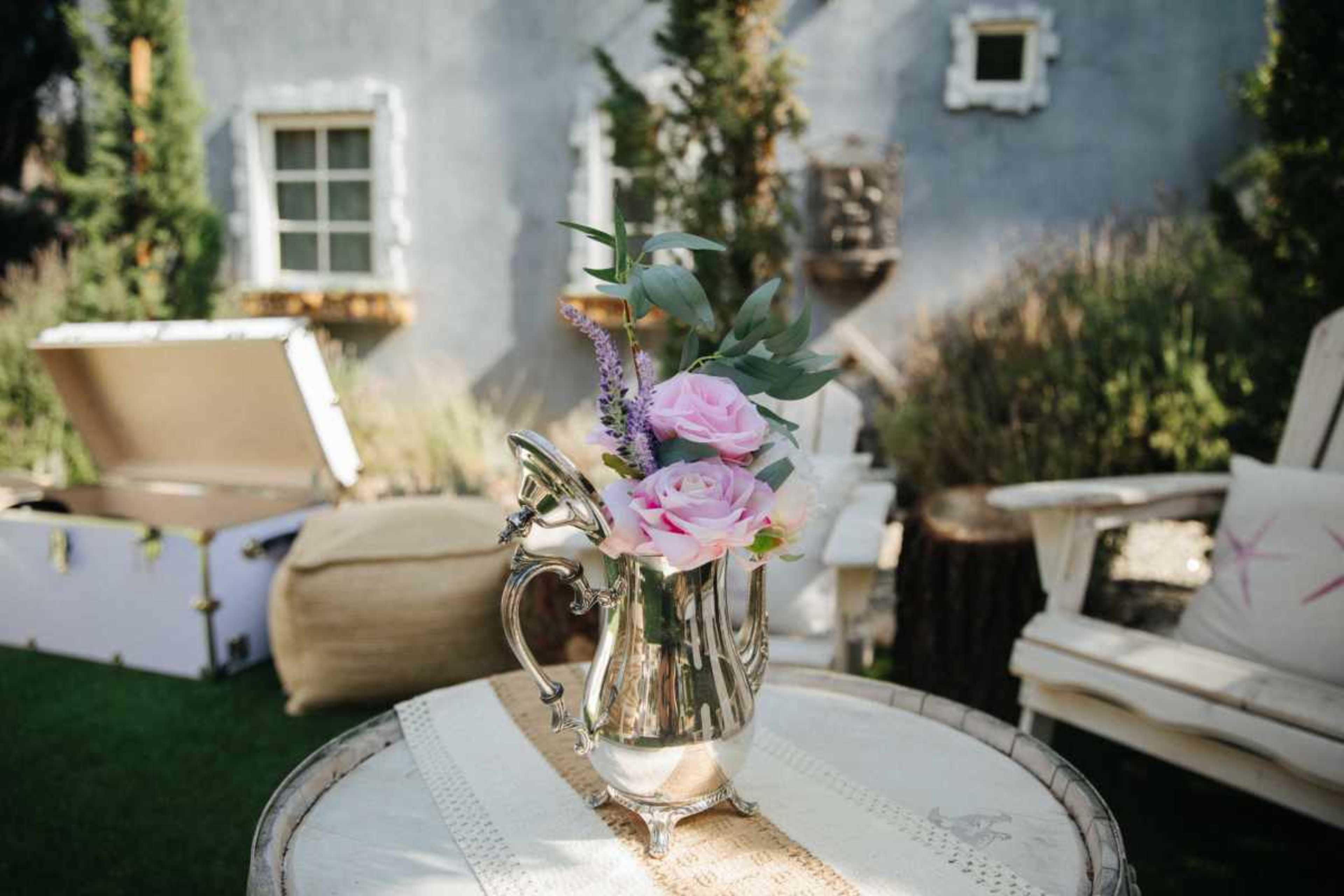 A silver pitcher filled with pink roses and greenery sits on a wooden table in a garden setting.