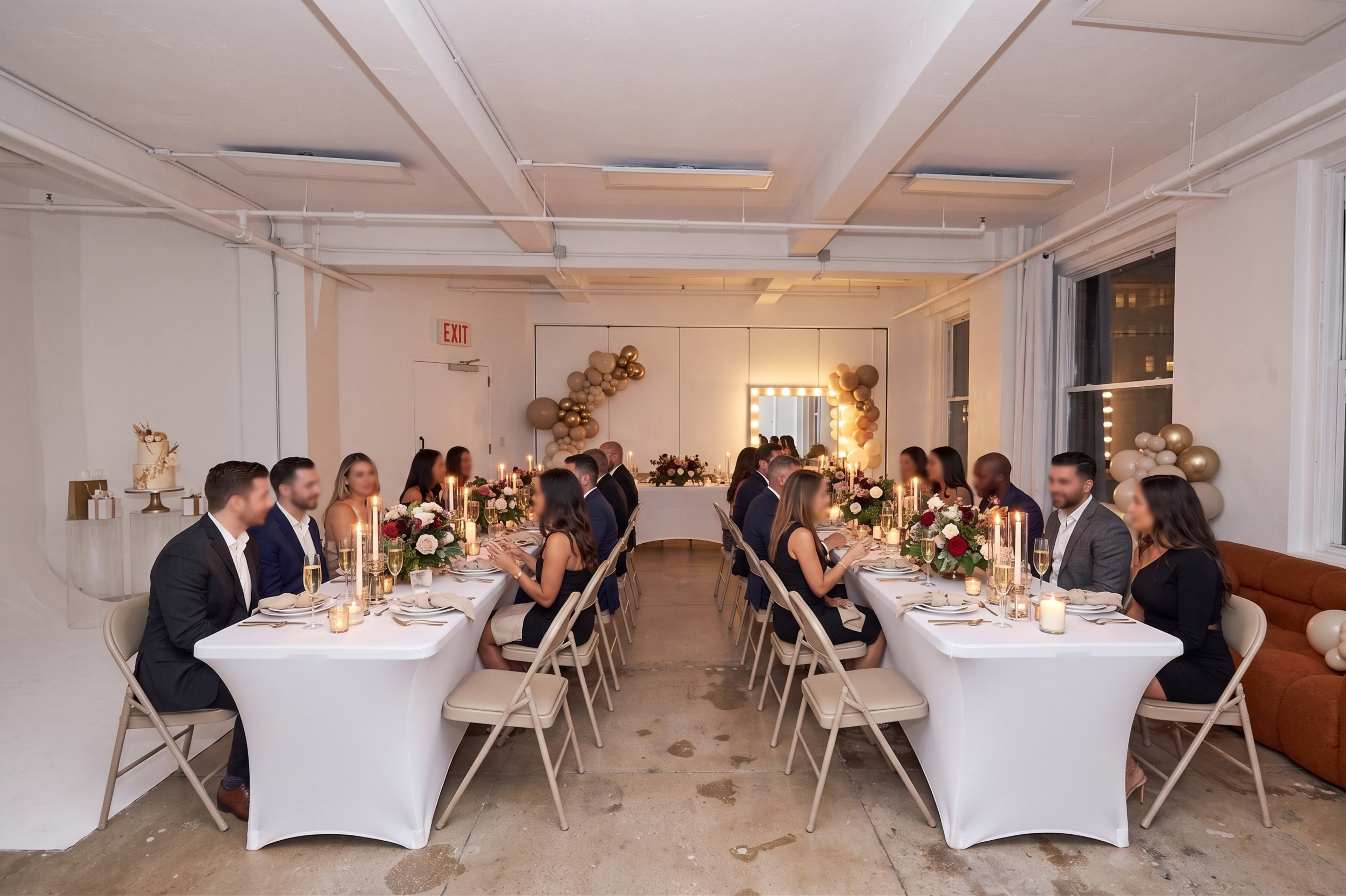 The image shows a formal dining setup with two long tables, decorated with flowers and candles, where a group of guests is seated, facing each other in a well-lit room.