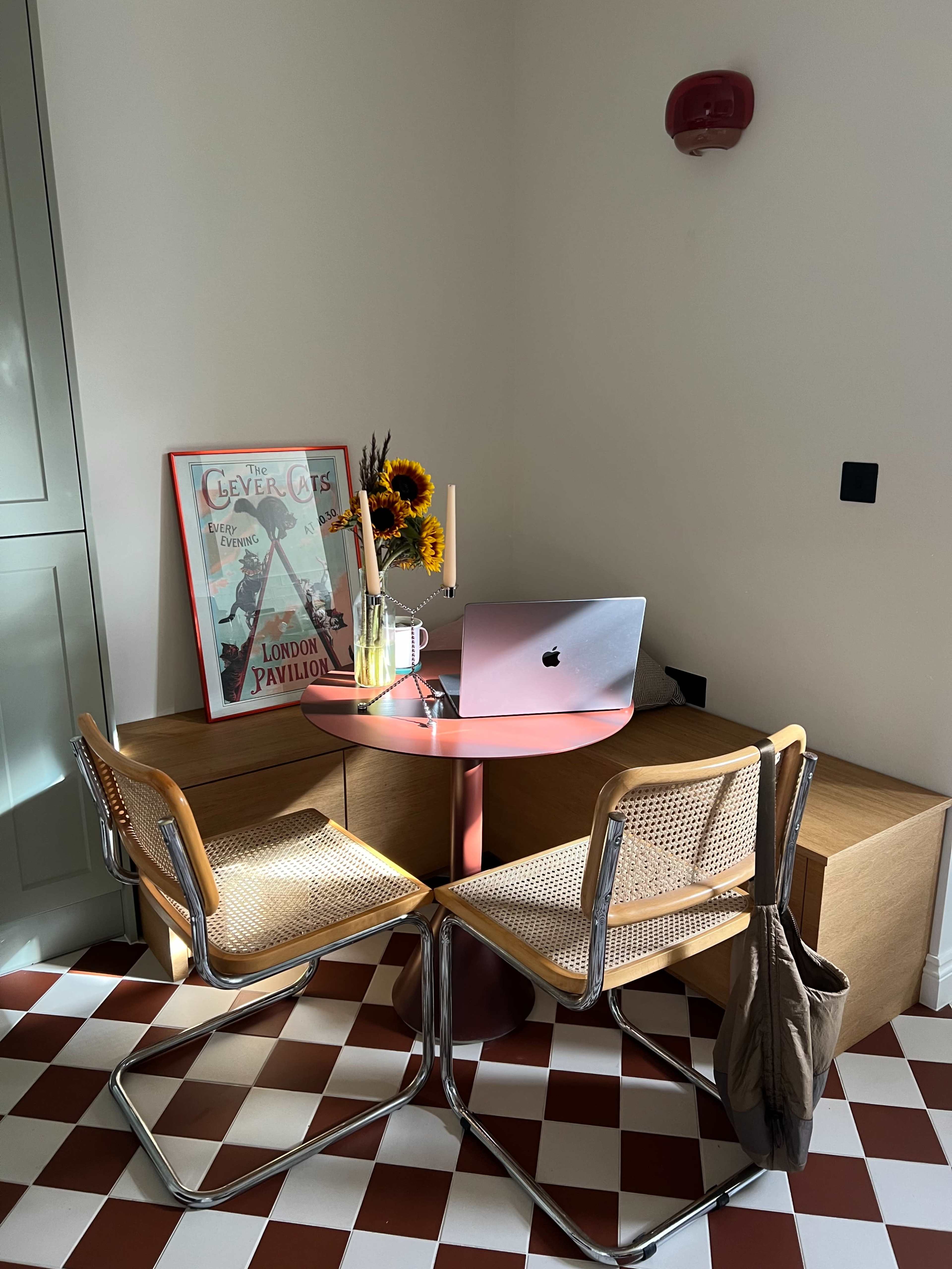 The image shows a small dining area with a round pink table, two chairs, a framed poster on the wall, a laptop on the table, and a vase of sunflowers.