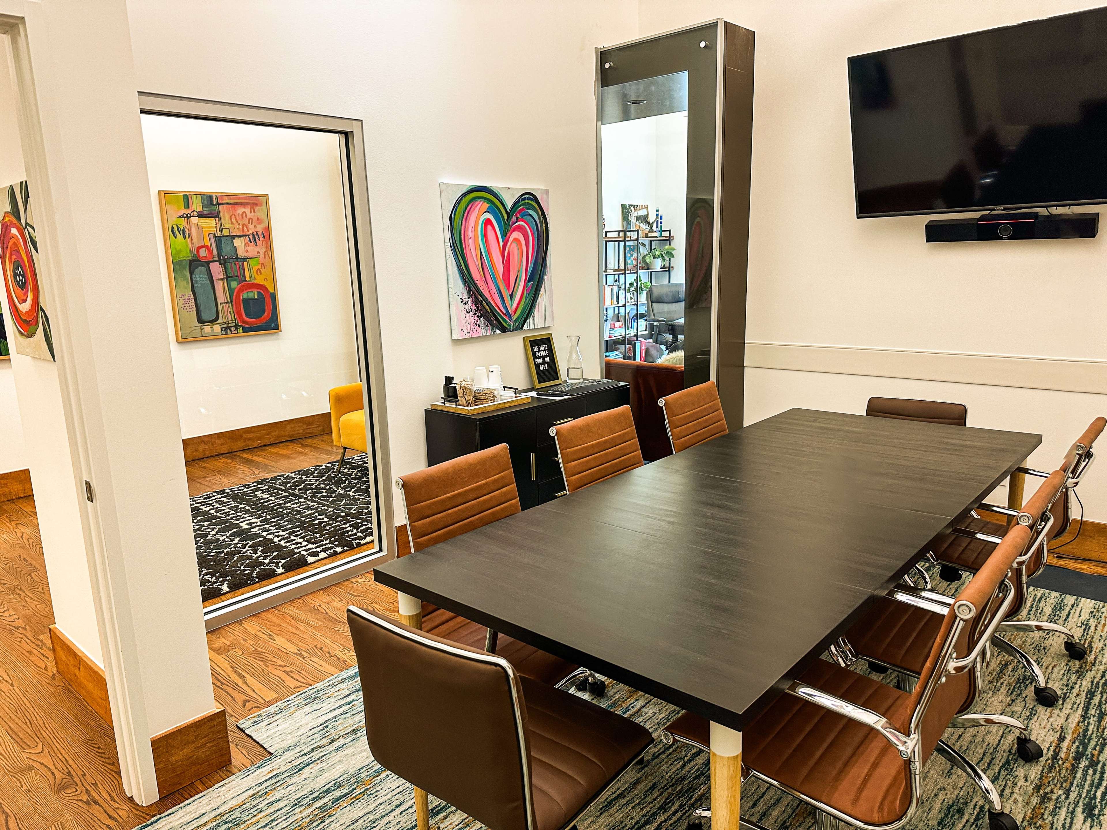 The image shows a modern conference room with a large black table surrounded by brown chairs, featuring colorful artwork on the walls and a glass-walled office visible through an open doorway.
