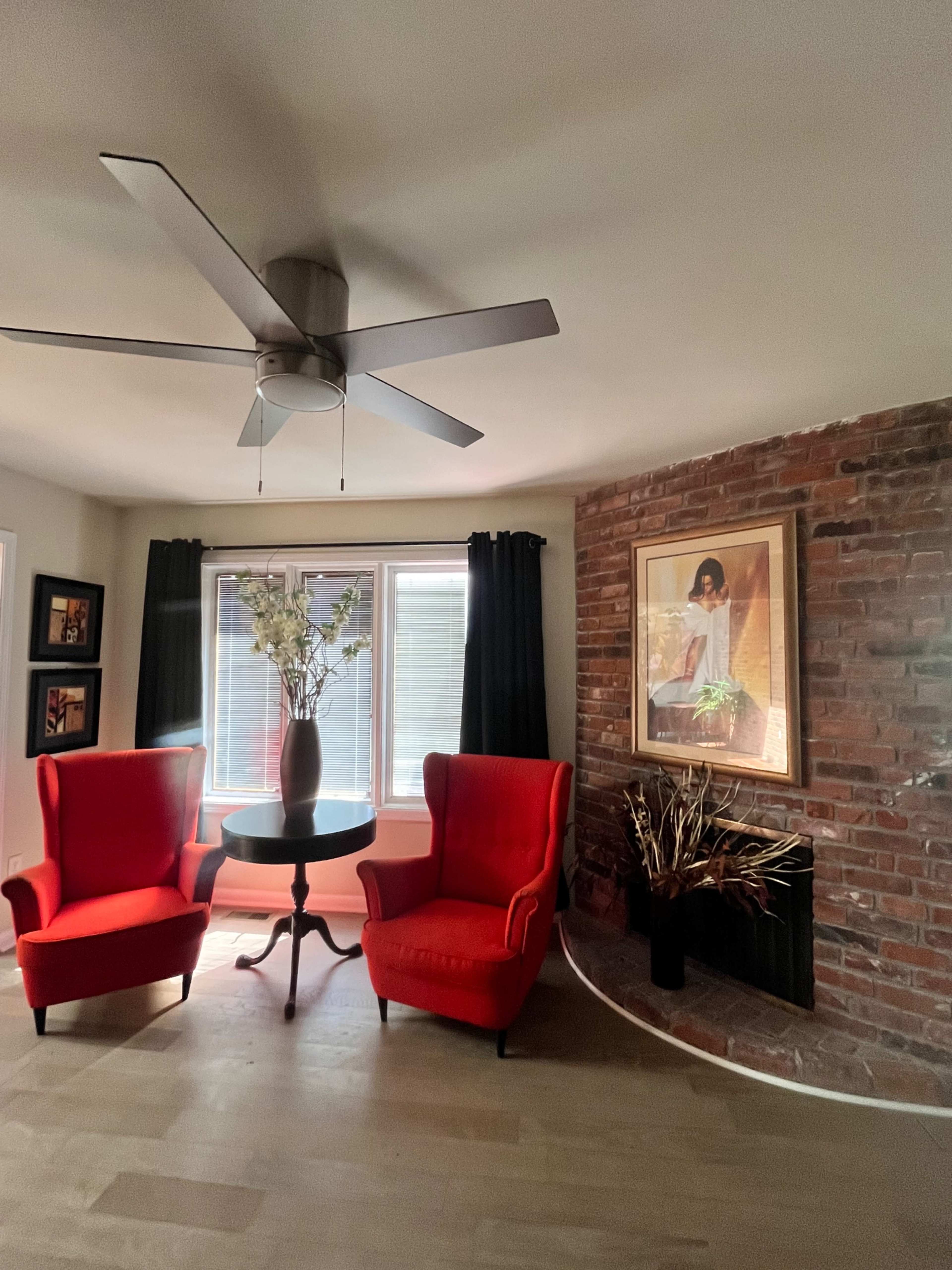 Two red armchairs are positioned around a small circular table in a living room with a brick accent wall and large windows covered by black curtains.