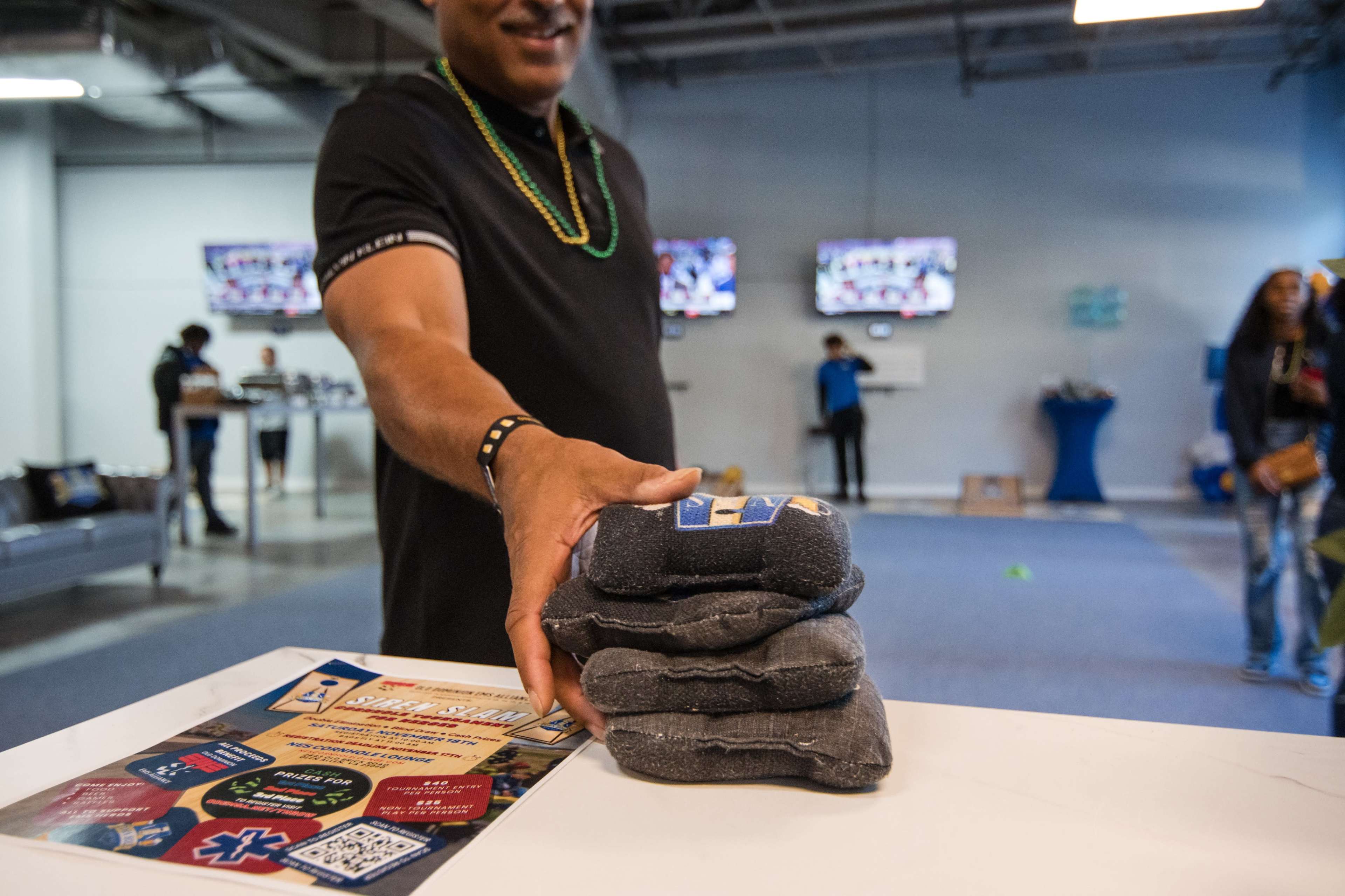 A person in a black shirt extends their hand to offer a stack of four gray bags on a table in a room with multiple screens displaying content in the background.
